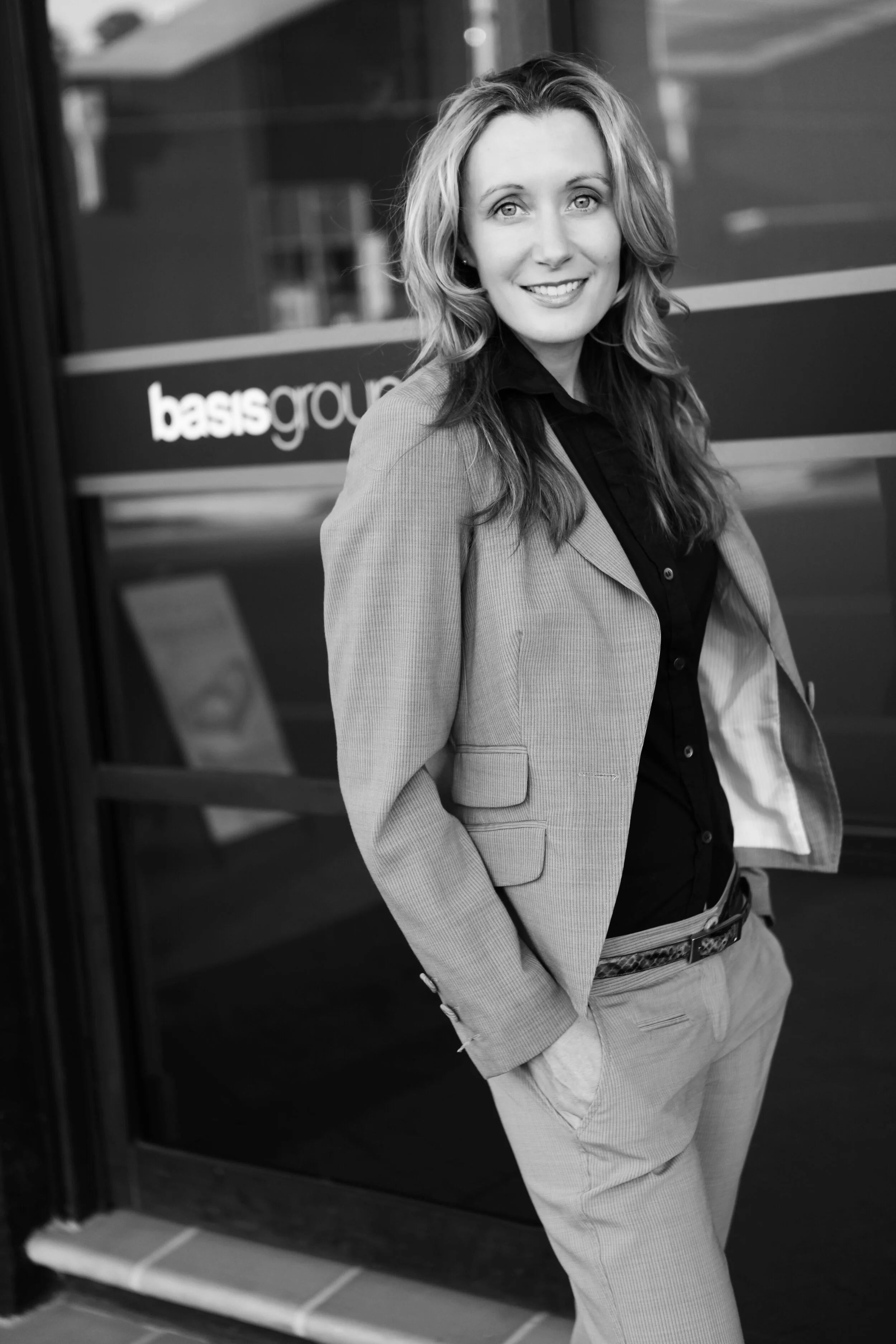 Black and white photo of a woman dressed in business attire standing outside a building with a sign that reads 'bassment.' She has long, wavy hair and is smiling, with one hand in her pocket.