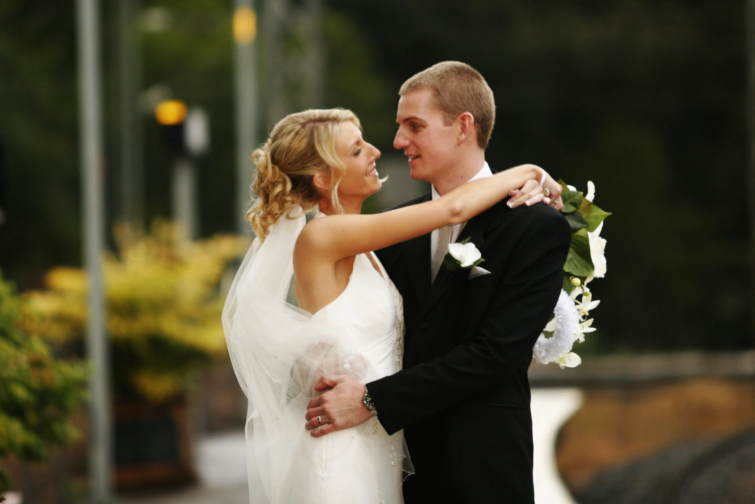 A bride and groom share a dance outdoors on their wedding day, smiling at each other. The bride wears a white dress with a veil, and the groom is in a black tuxedo with a white boutonniere.