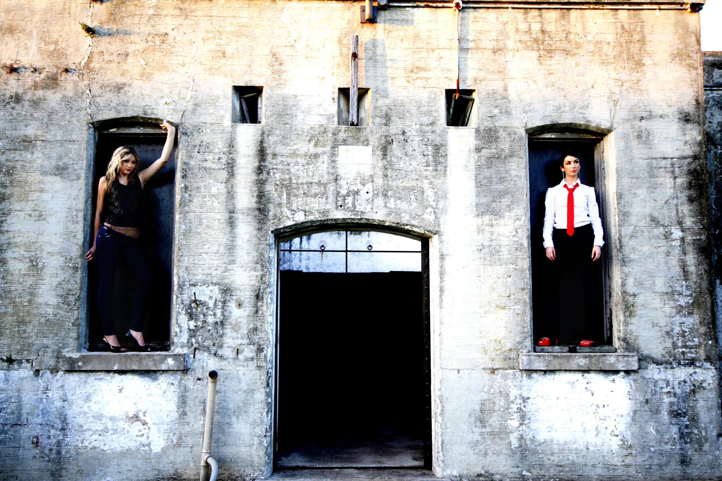 Two women standing in open window frames of a weathered concrete building, one on the left in black clothing and high heels, the other on the right in white shirt, red tie, and sandals.