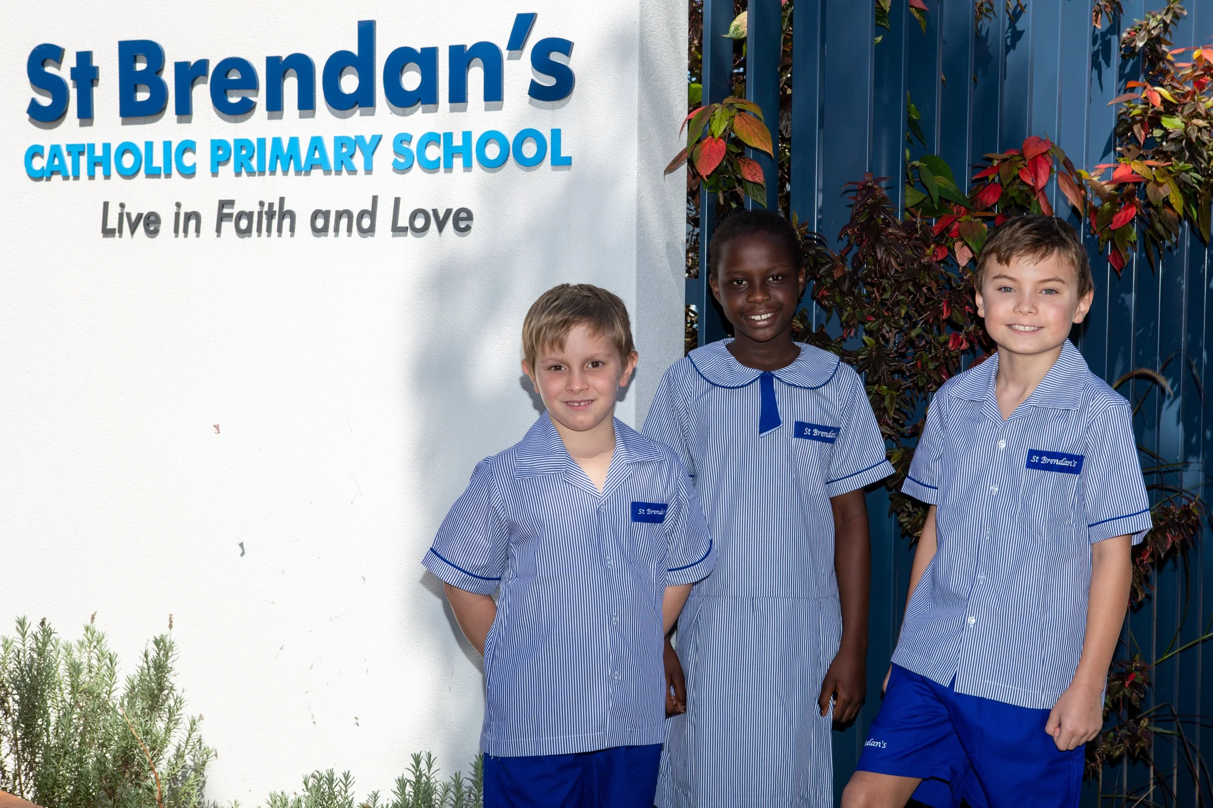 Three children in blue and white school uniforms stand next to a sign that reads 'St Brendan's Catholic Primary School, Live in Faith and Love' outside a school building, with greenery and a blue fence in the background.