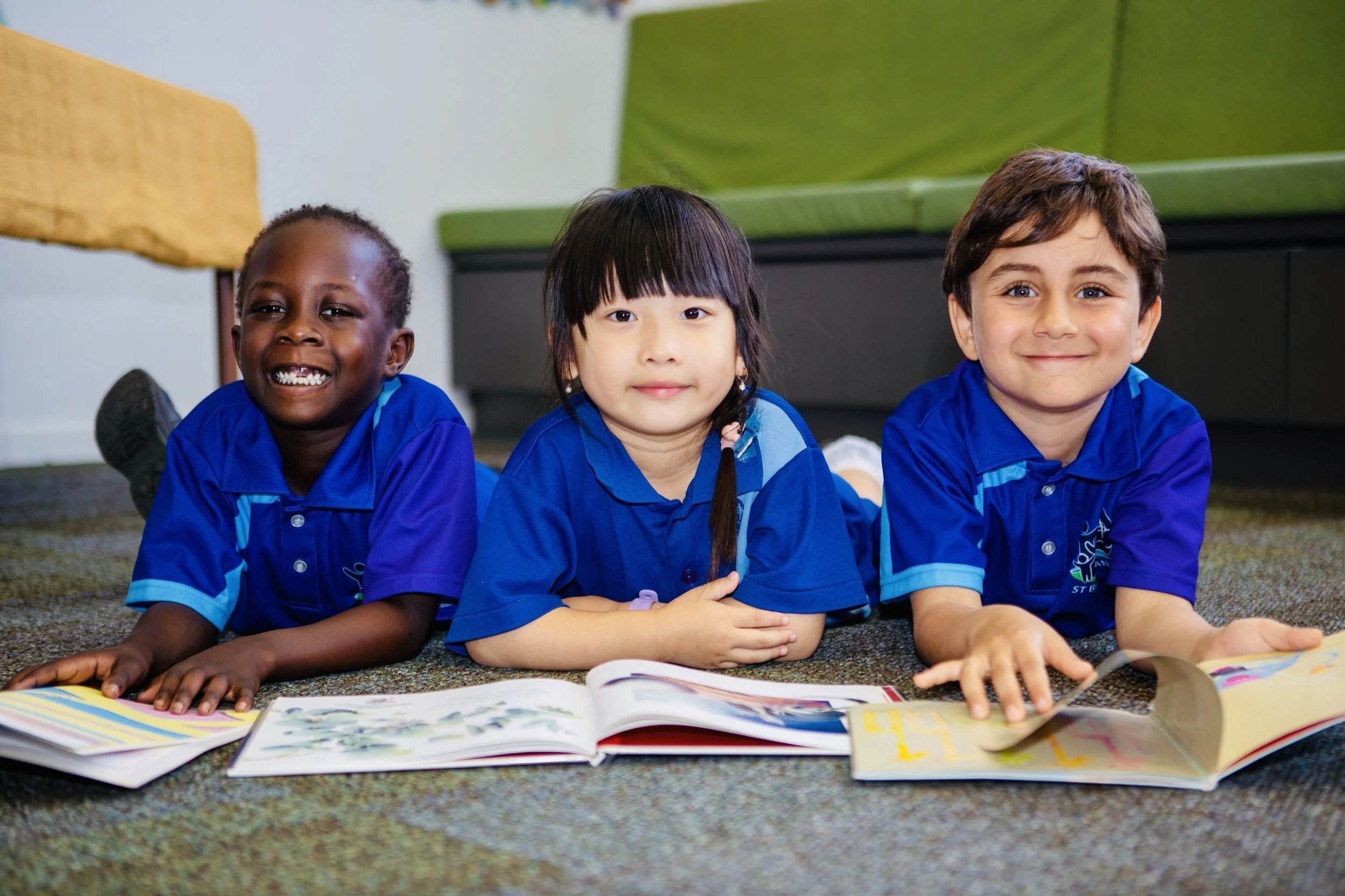 Three children lying on their stomachs on a carpeted floor, looking at open books, wearing matching blue school uniforms.