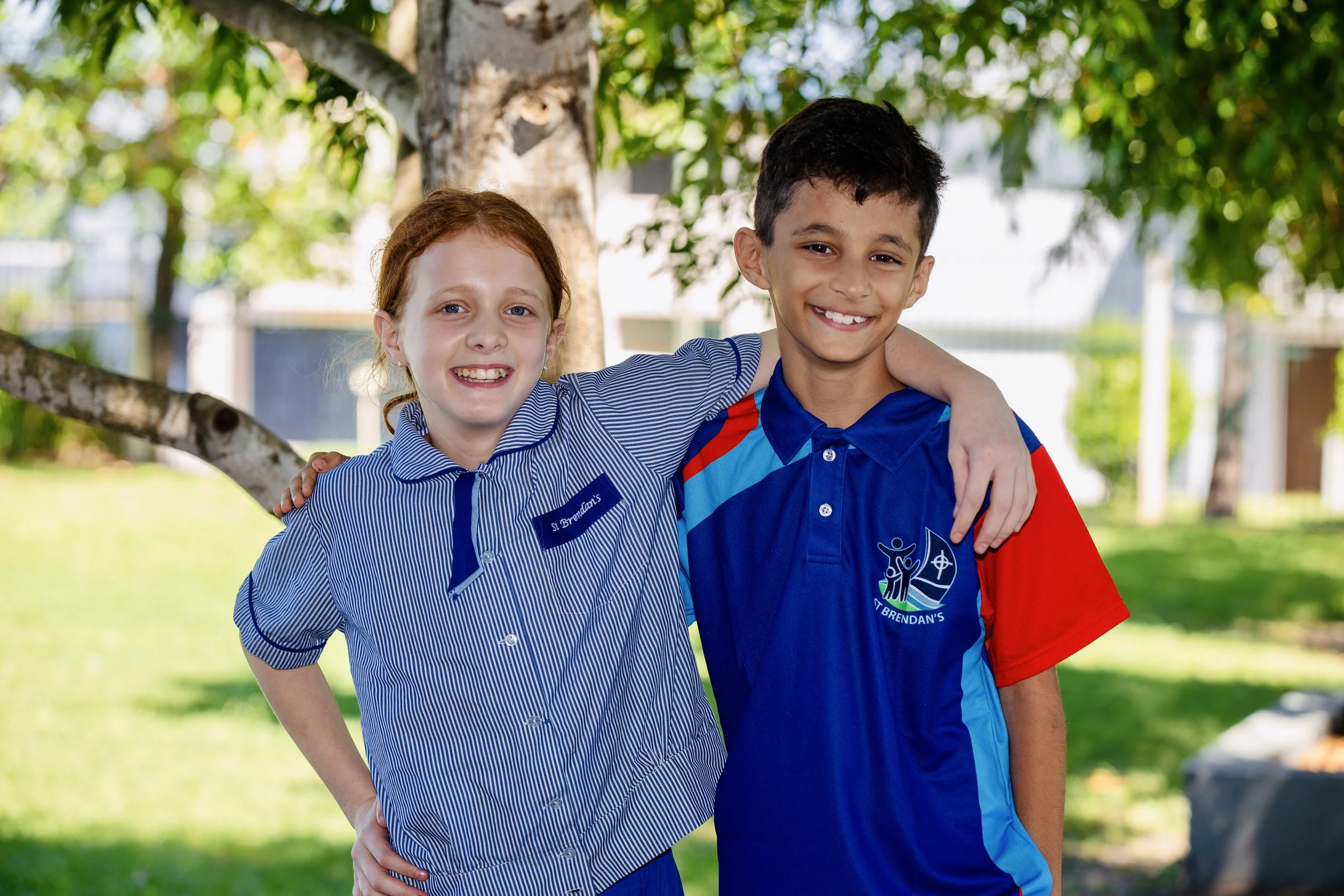 Two schoolchildren, a girl with red hair and a boy with dark hair, smiling, standing outdoors with their arms around each other. The girl is wearing a blue striped school uniform with a badge labeled 'St Brendan's' and the boy is in a blue and red sp