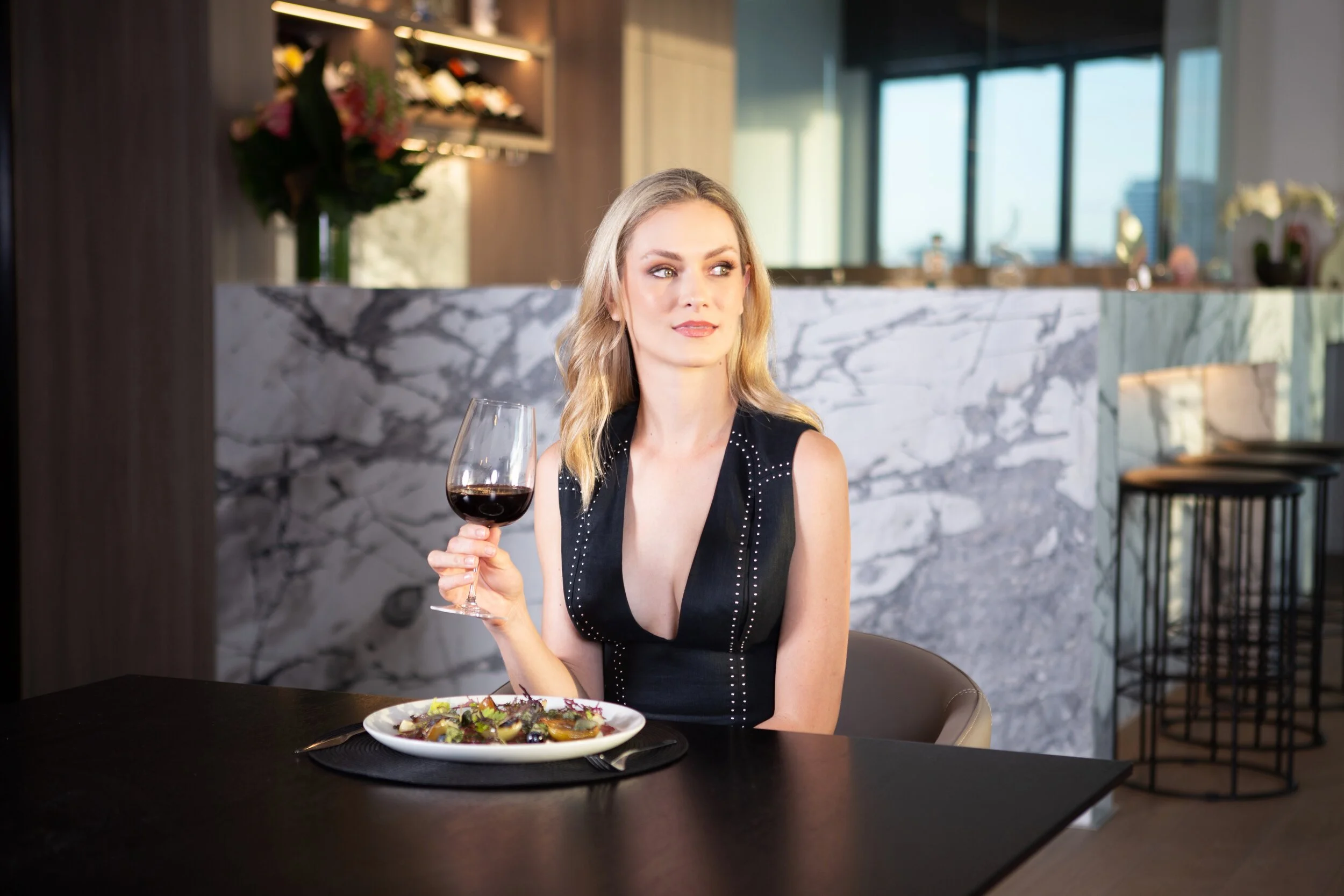 A woman with blonde hair in a black dress with silver accents sitting at a table in a restaurant, holding a glass of red wine next to a plate of mixed salad.