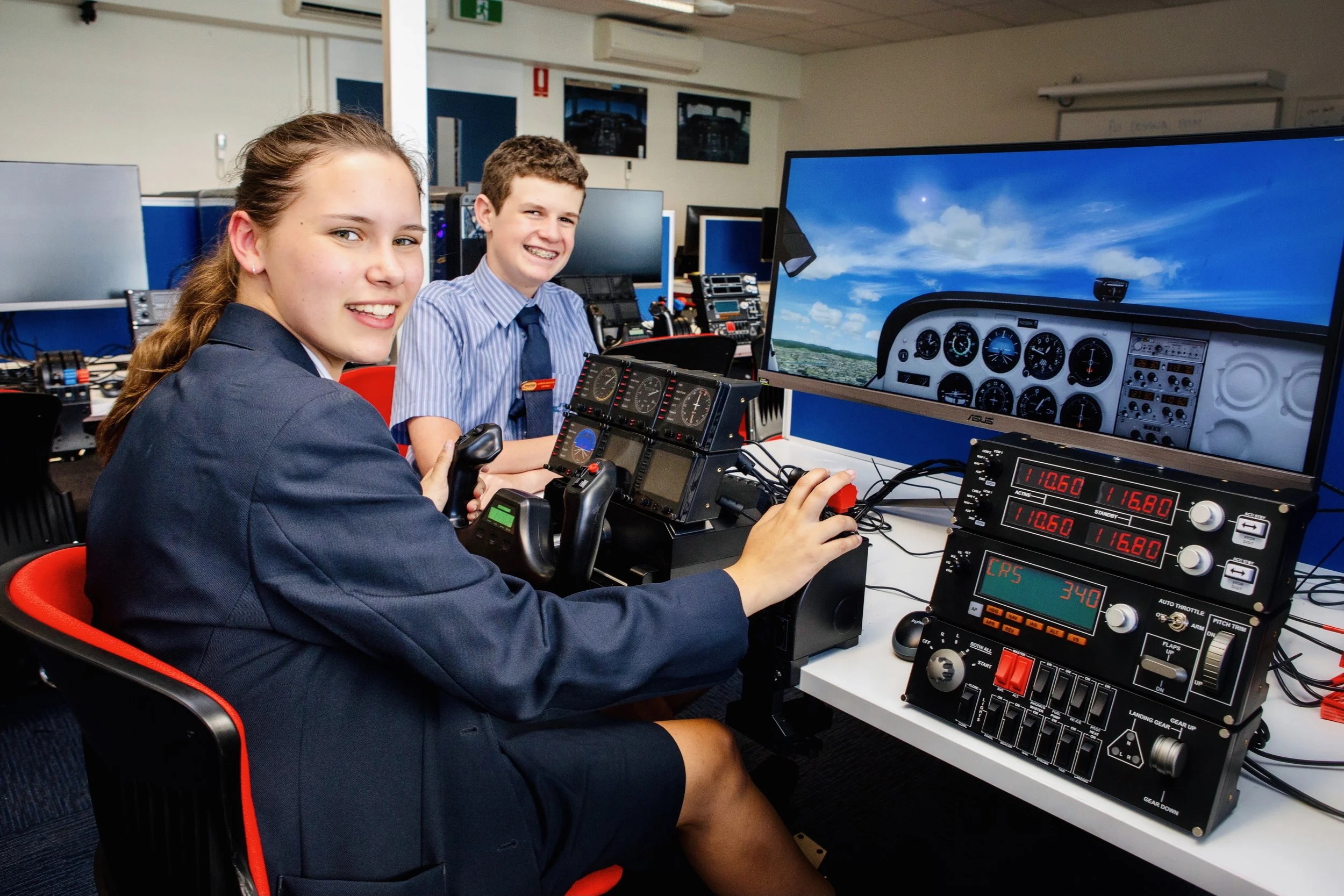Two students, a girl and a boy, sitting at a desk with airplane simulation equipment, including a joystick and instruments, in a classroom setting. The girl is operating the controls, looking at the camera with a smile. A large screen displays a cock