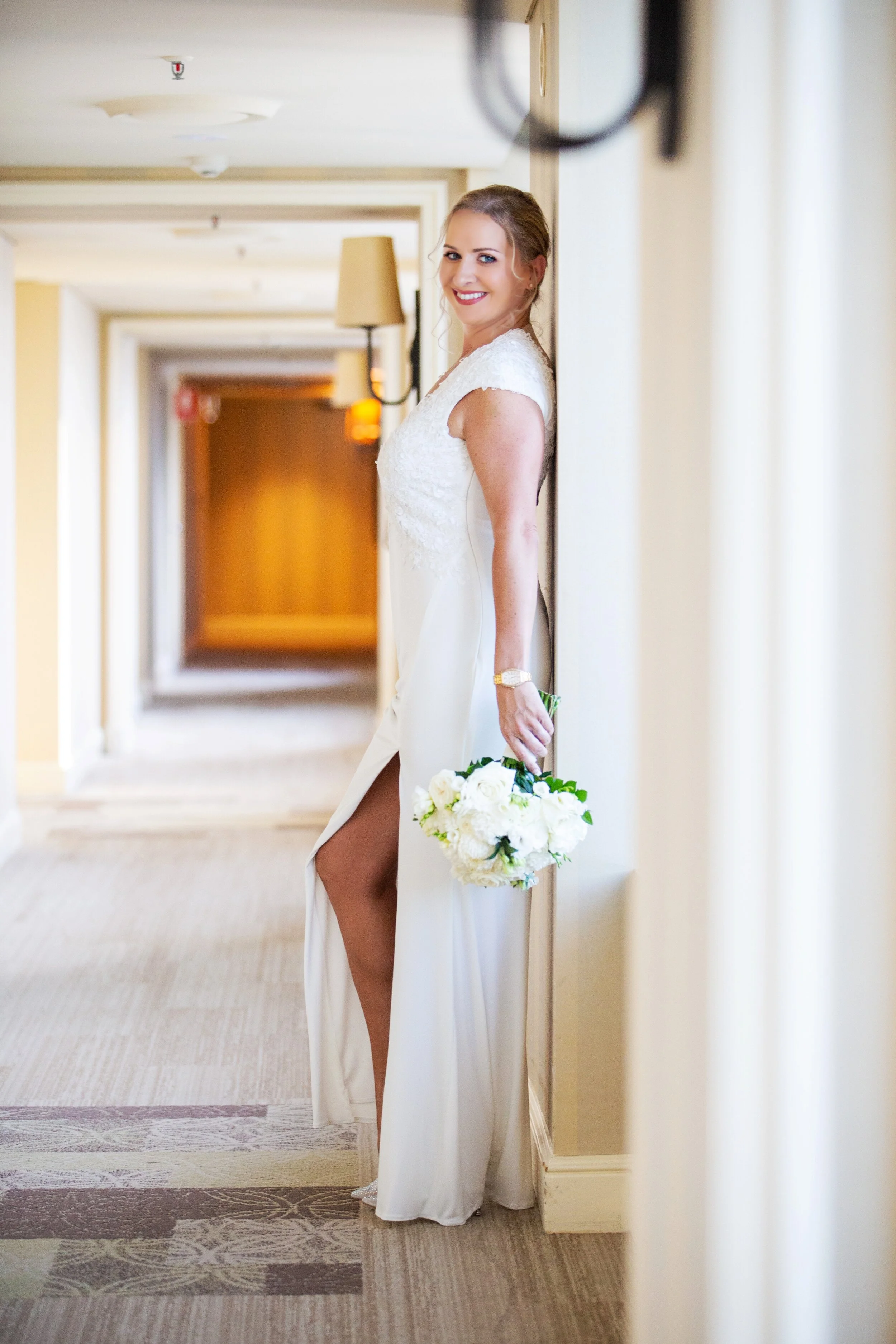 A woman in a white wedding dress with a slit, holding a bouquet of white flowers, smiling while standing in a hallway.