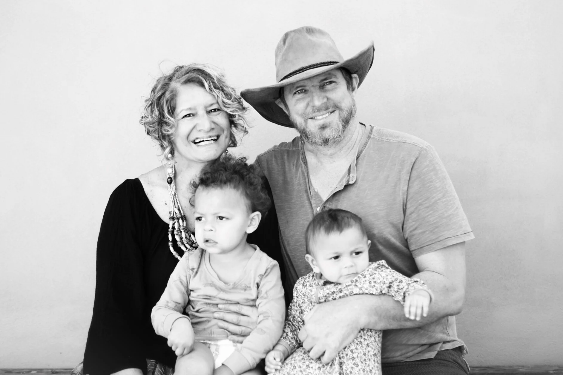 A smiling woman with curly hair, a man with a beard wearing a hat, and two young children sitting together against a plain background.