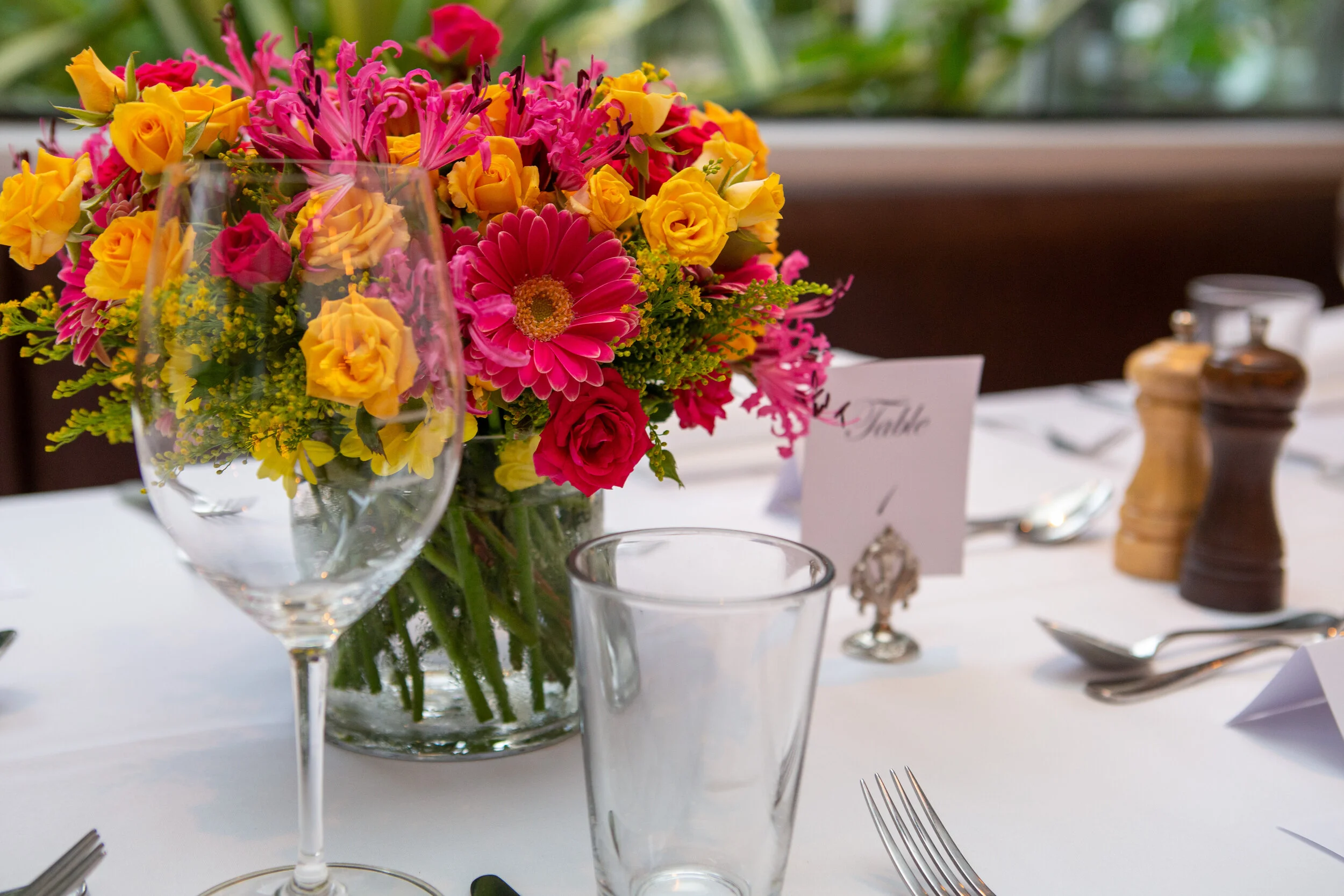 A dining table set with a colorful bouquet of pink, yellow, and orange flowers, surrounded by glassware, silverware, and salt and pepper shakers, with a white tablecloth.