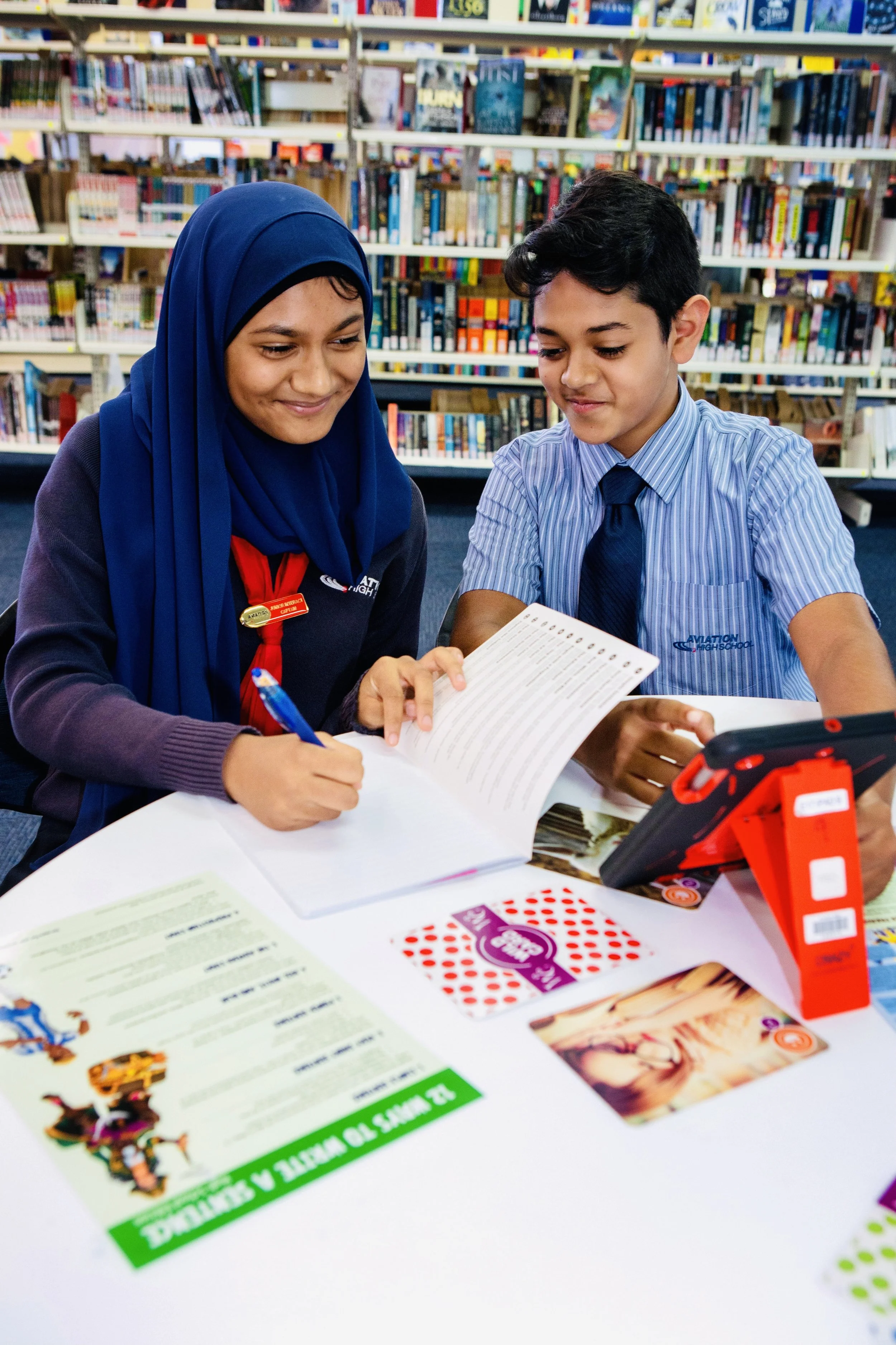 A young girl and boy in school uniforms sitting at a table in a library, reviewing a page in a book together.