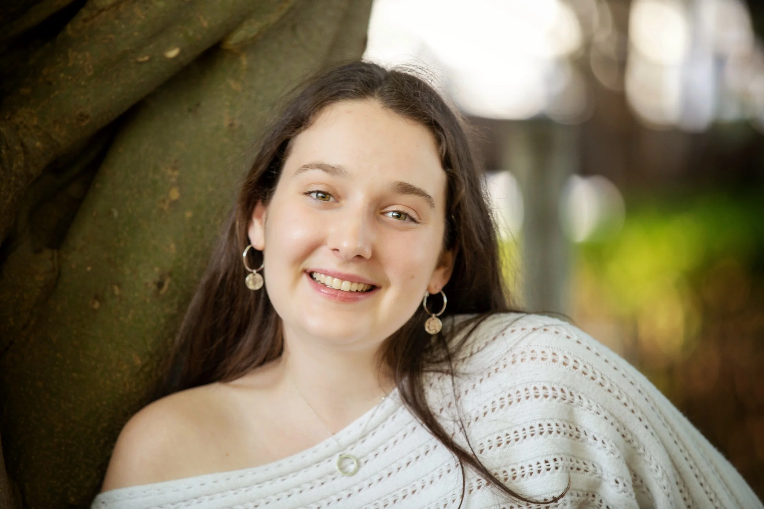 A young woman with long brown hair smiling and leaning against a tree outdoors.