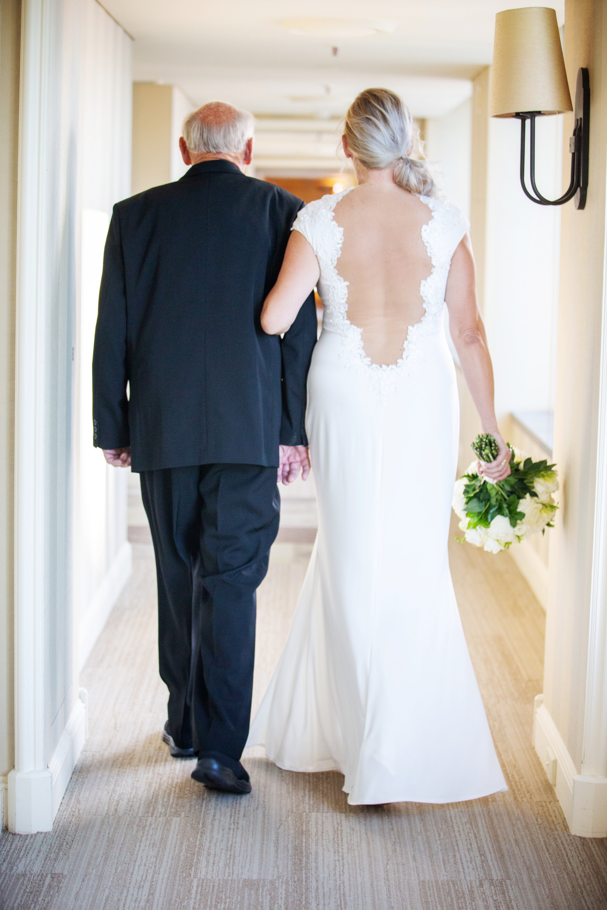 A bride in a white wedding dress walking arm-in-arm with an older man, likely her father, down a hallway. The bride is holding a bouquet of white flowers and has a lace-backed dress.