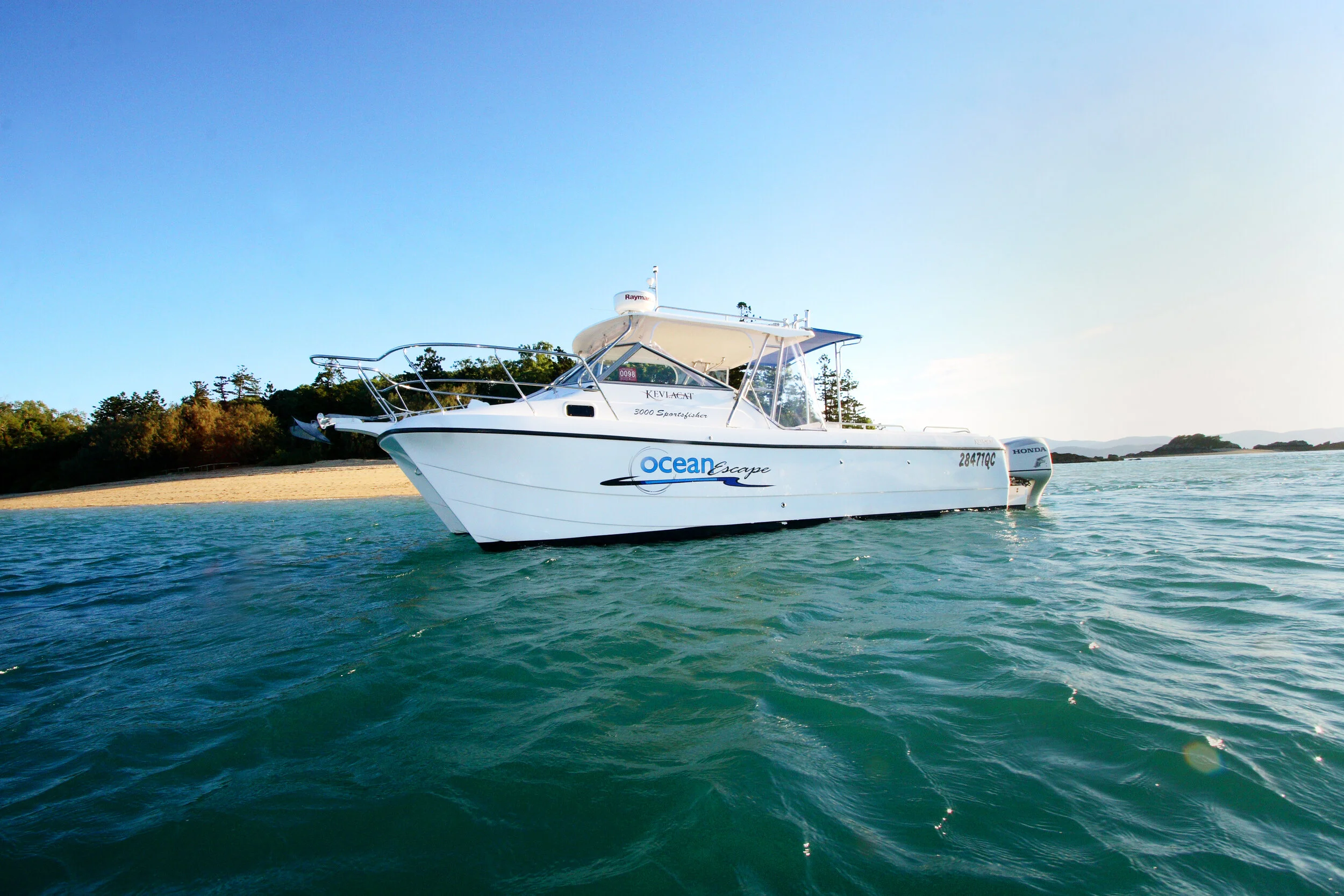A white motorboat named Ocean Escape floating on calm water near a sandy beach with trees in the background.
