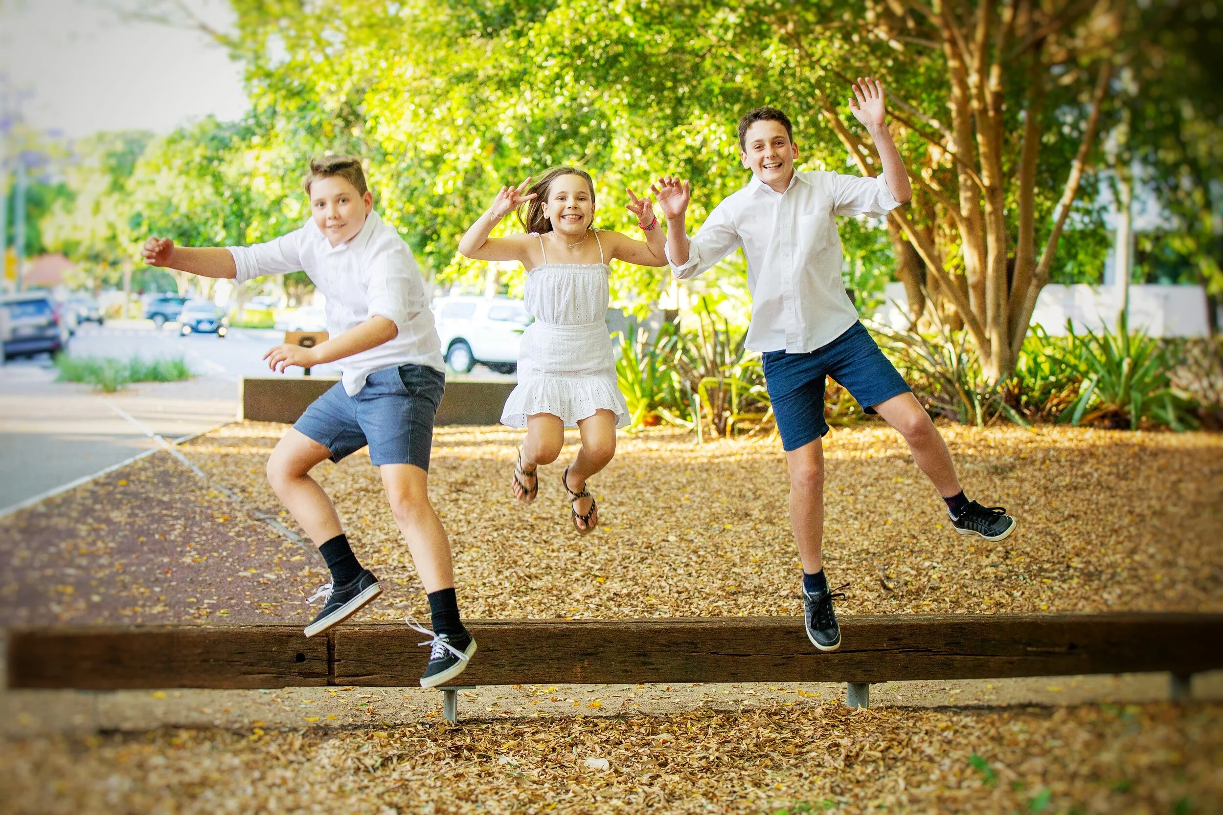 Three children jumping and playing on a wooden beam in a park with trees and parked cars in the background.