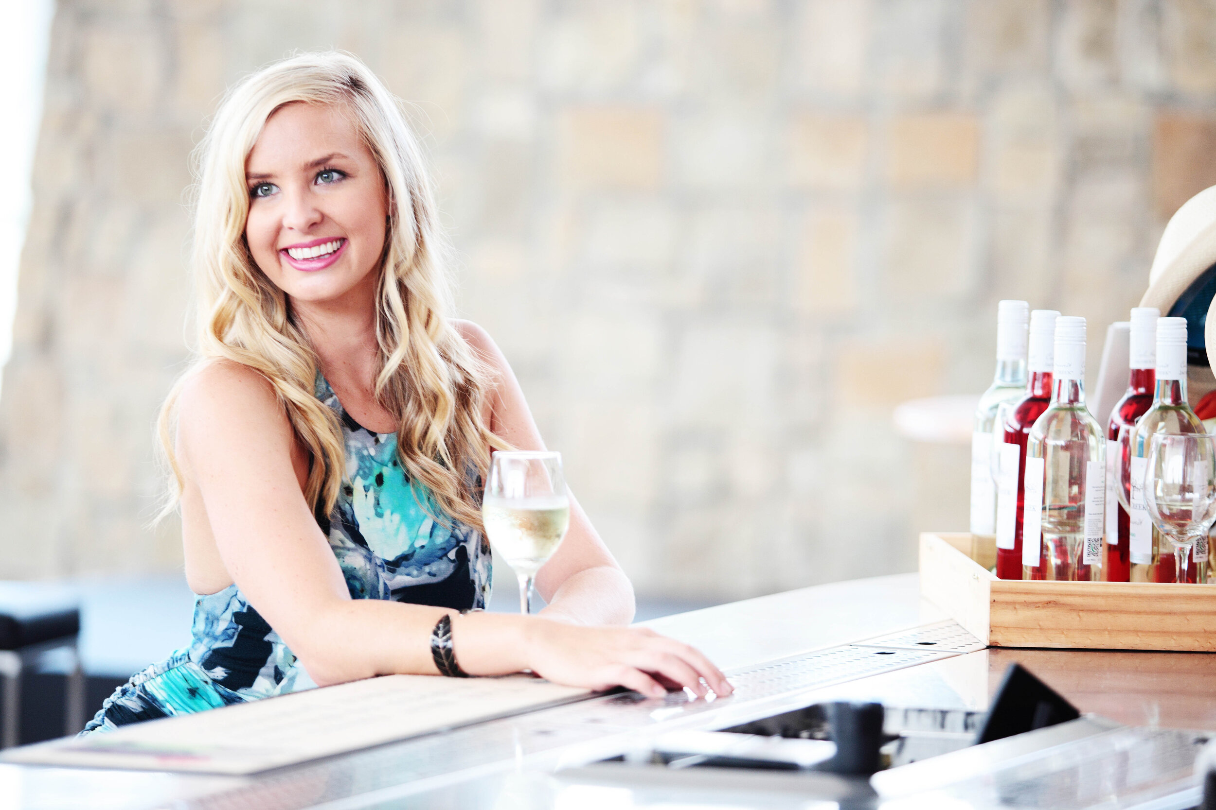 A smiling woman with long blonde hair sitting at a bar with a glass of white wine in front of her. Bottles of alcohol are in a wooden tray on the bar.
