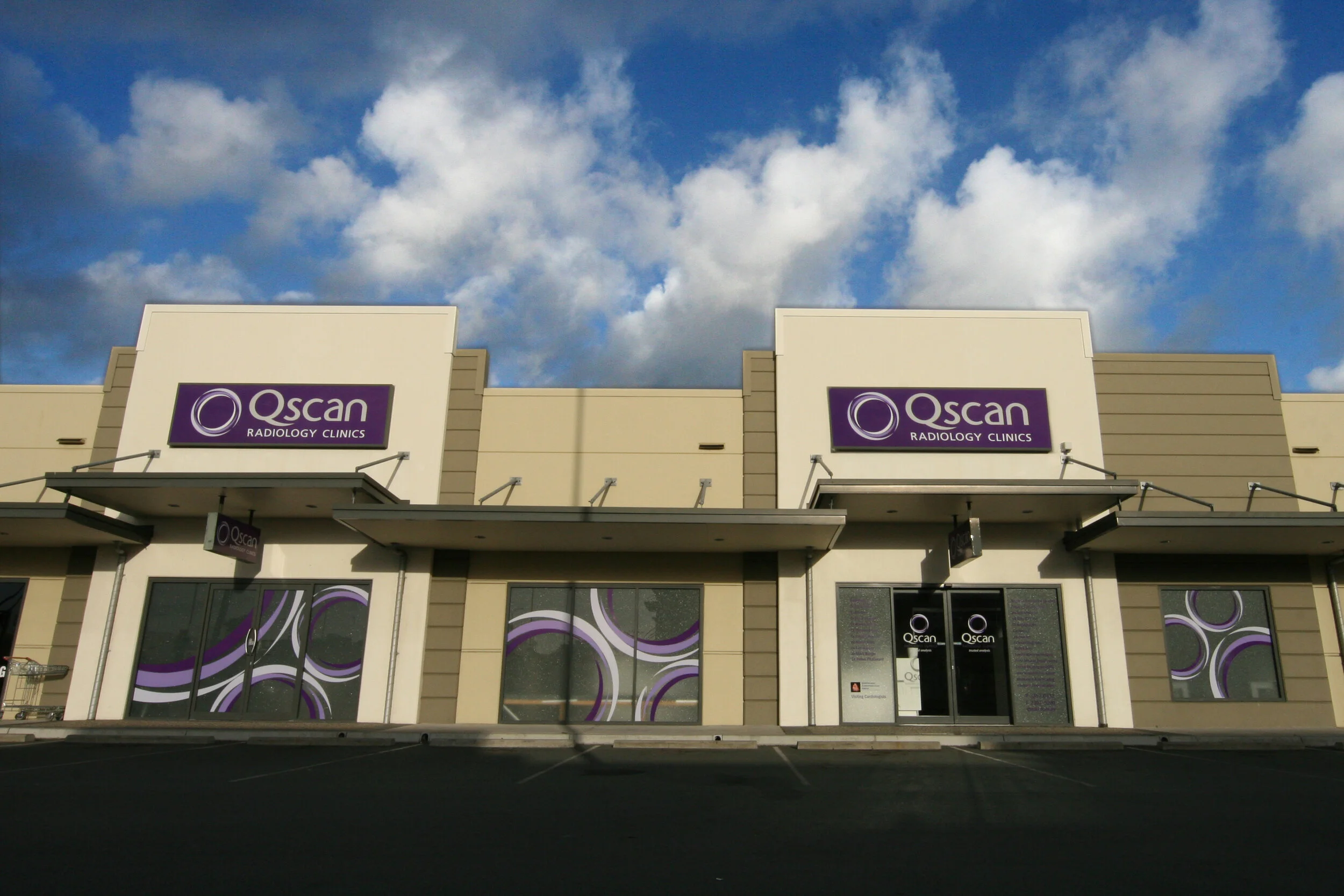 Qscan Radiology Clinics building with purple and white signs under a partly cloudy blue sky, featuring large windows with purple and white circular designs.