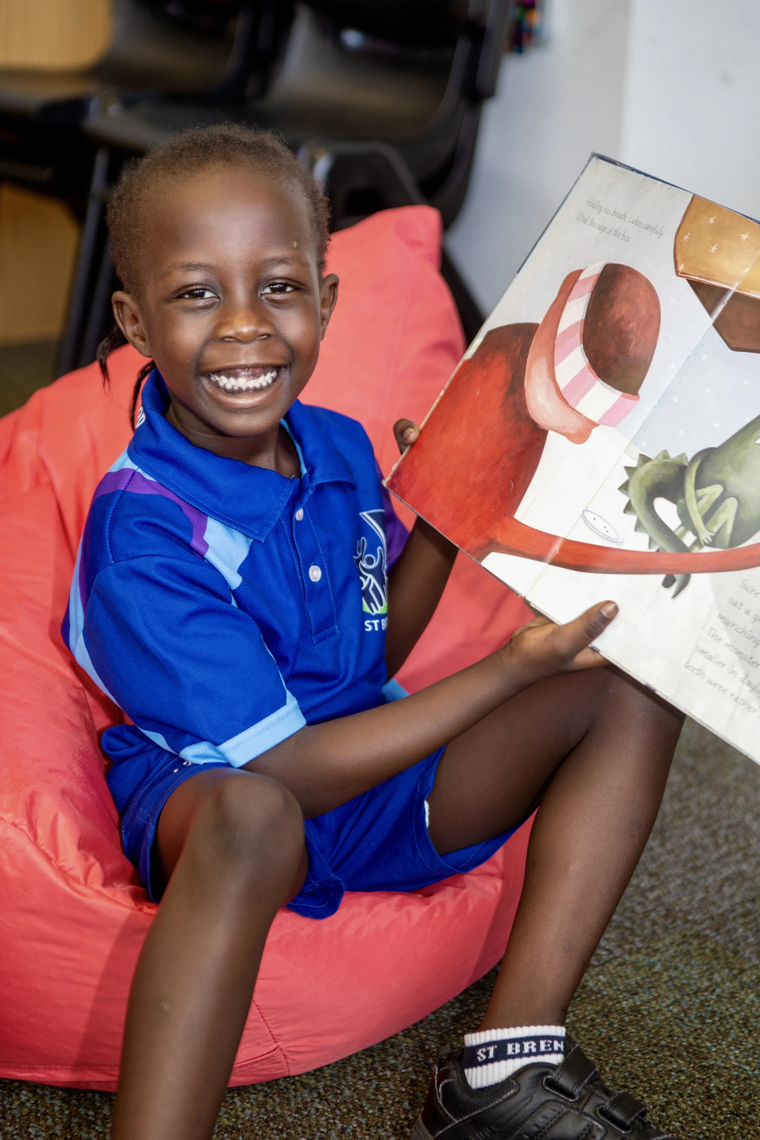 A young boy sits on a red bean bag chair, smiling and reading a large illustrated book with colorful pictures of vegetables.