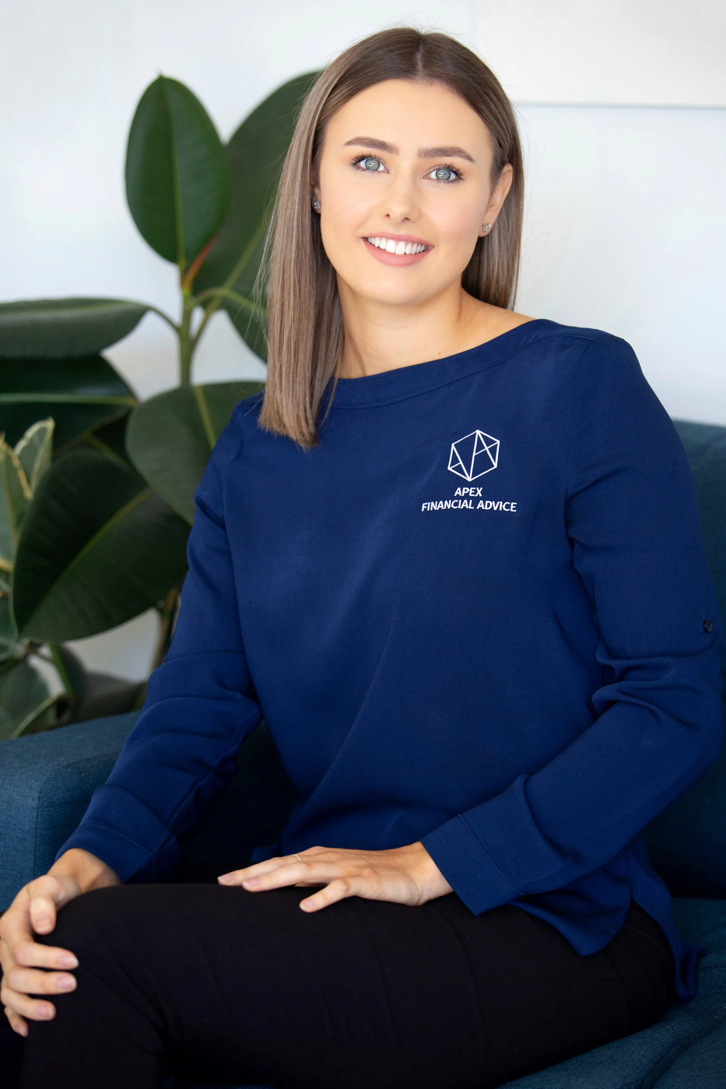 A woman with brown hair and blue eyes sitting on a blue couch, wearing a blue sweatshirt with the logo 'APEX FINANCIAL ADVICE' and a geometric design, smiling at the camera, with large green houseplants in the background.