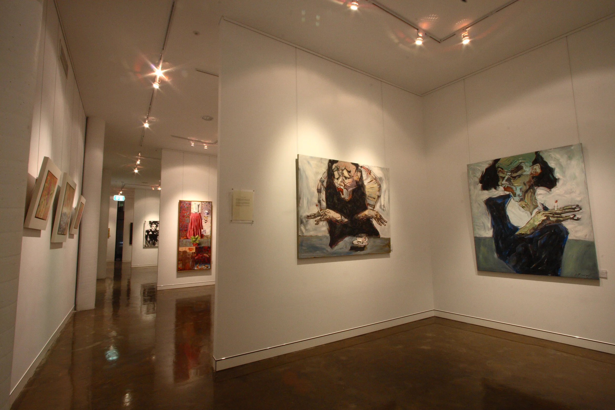 Art gallery featuring abstract and figurative paintings on white walls with wooden floors and ceiling spotlights.