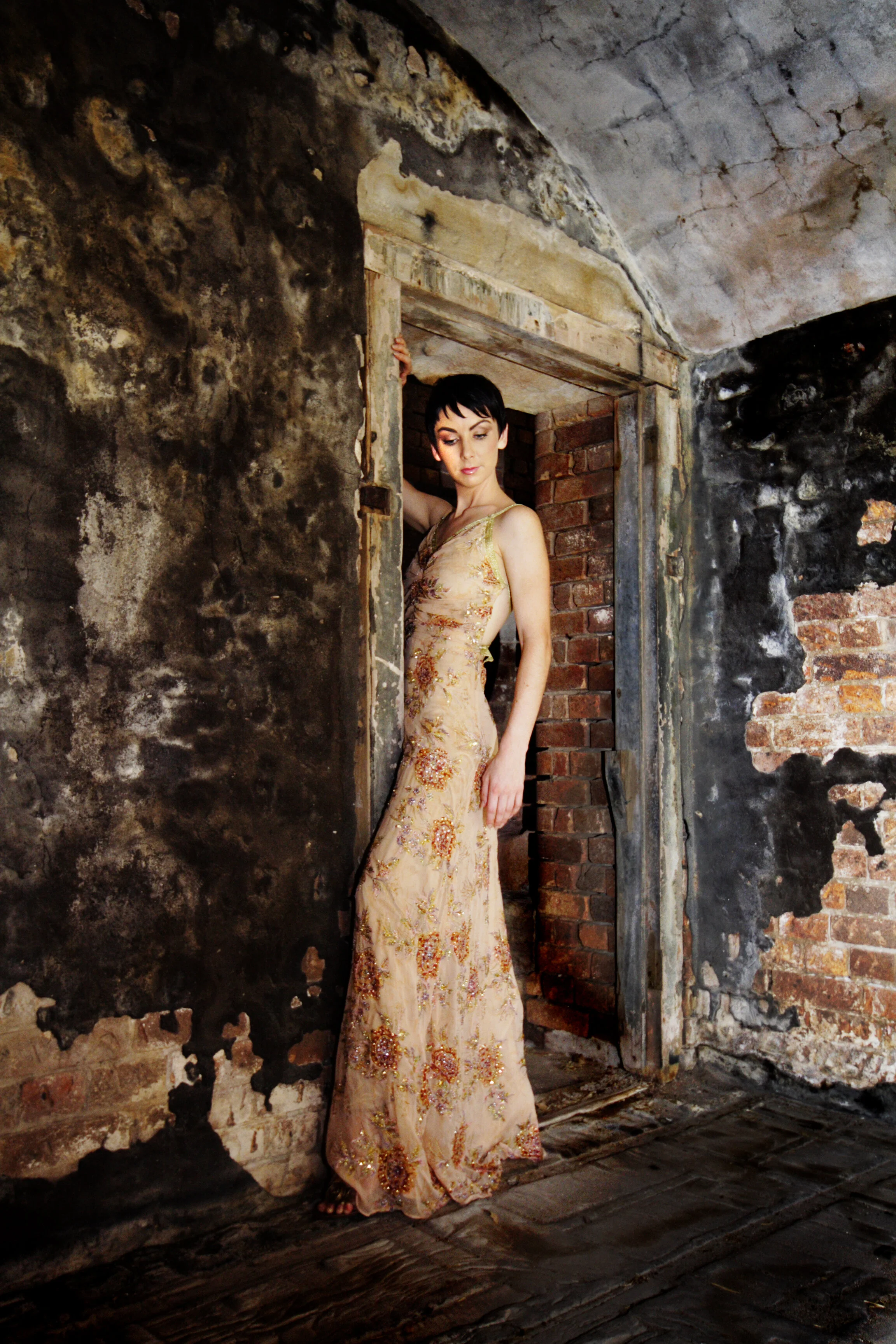 A woman with short black hair wearing a beige gown with floral embroidery poses in a rustic, unfinished room with brick and blackened walls.