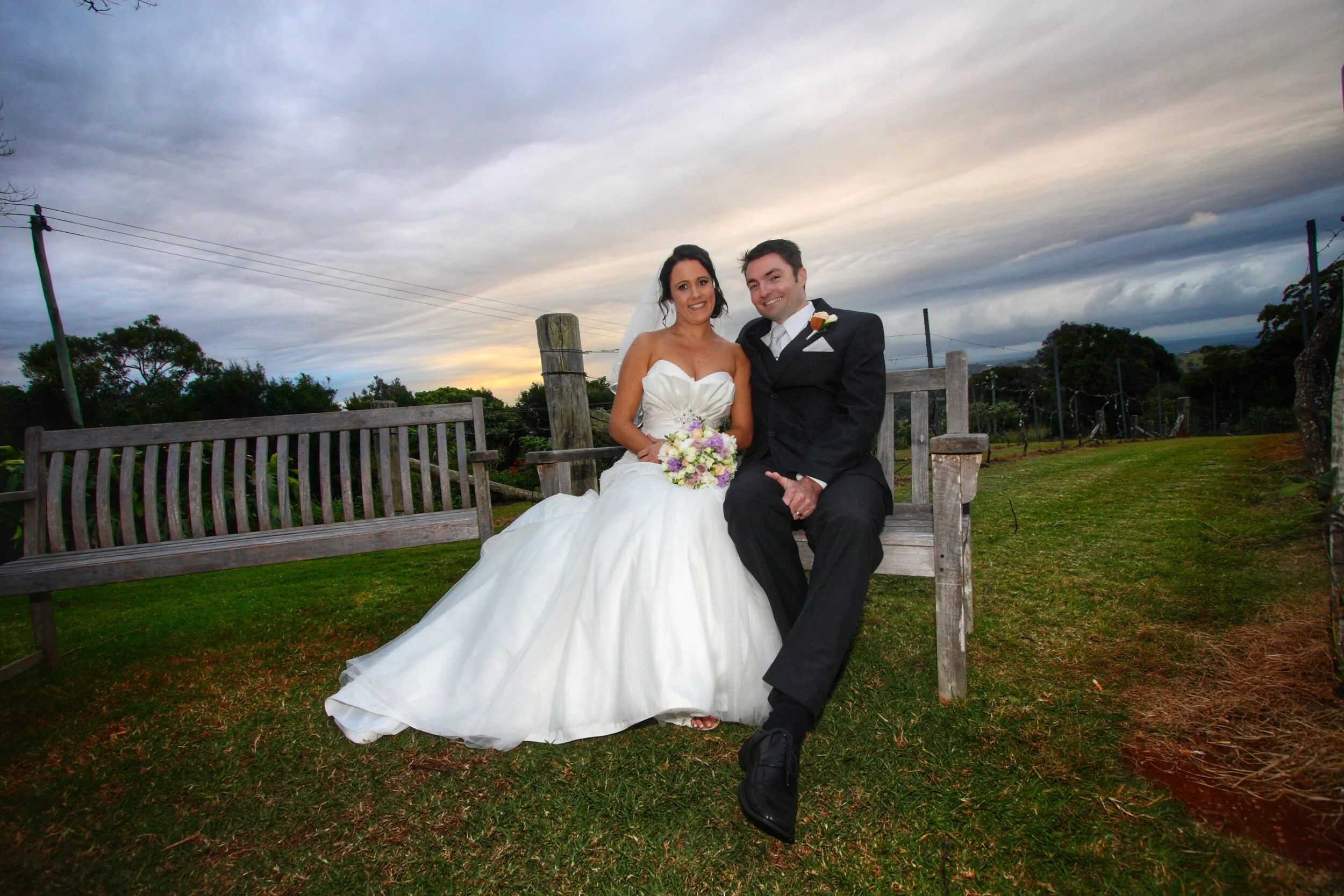 A bride and groom sitting on a park bench outdoors during sunset, smiling for a wedding photo. The bride is in a white wedding gown holding a bouquet, and the groom is in a black tuxedo with a boutonnière.