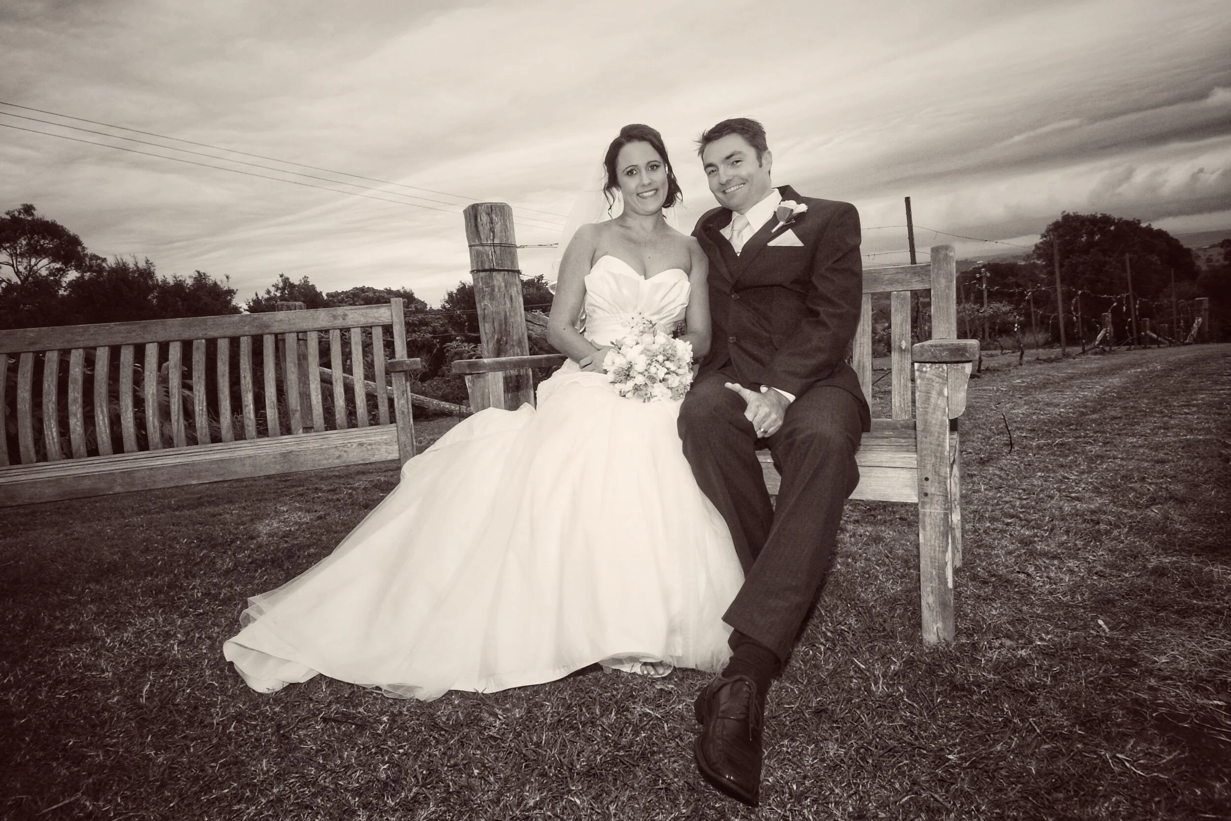 A bride and groom sitting on a wooden bench outside, dressed in wedding attire, smiling at the camera, with trees and cloudy sky in the background.