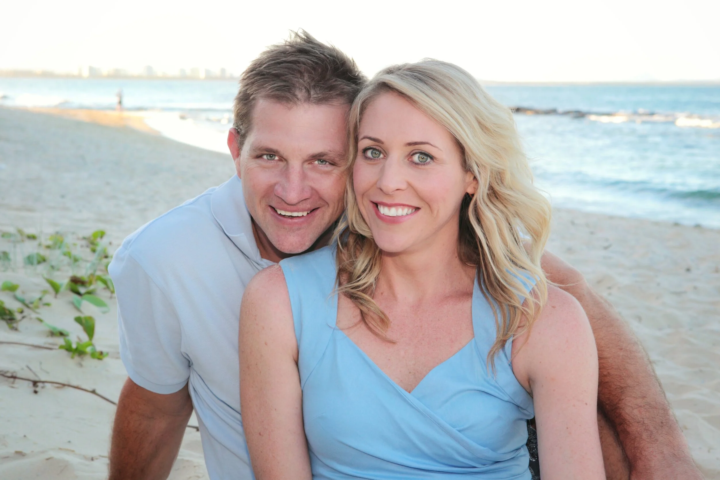 A happy couple sitting on the sandy beach with the ocean and city skyline in the background.
