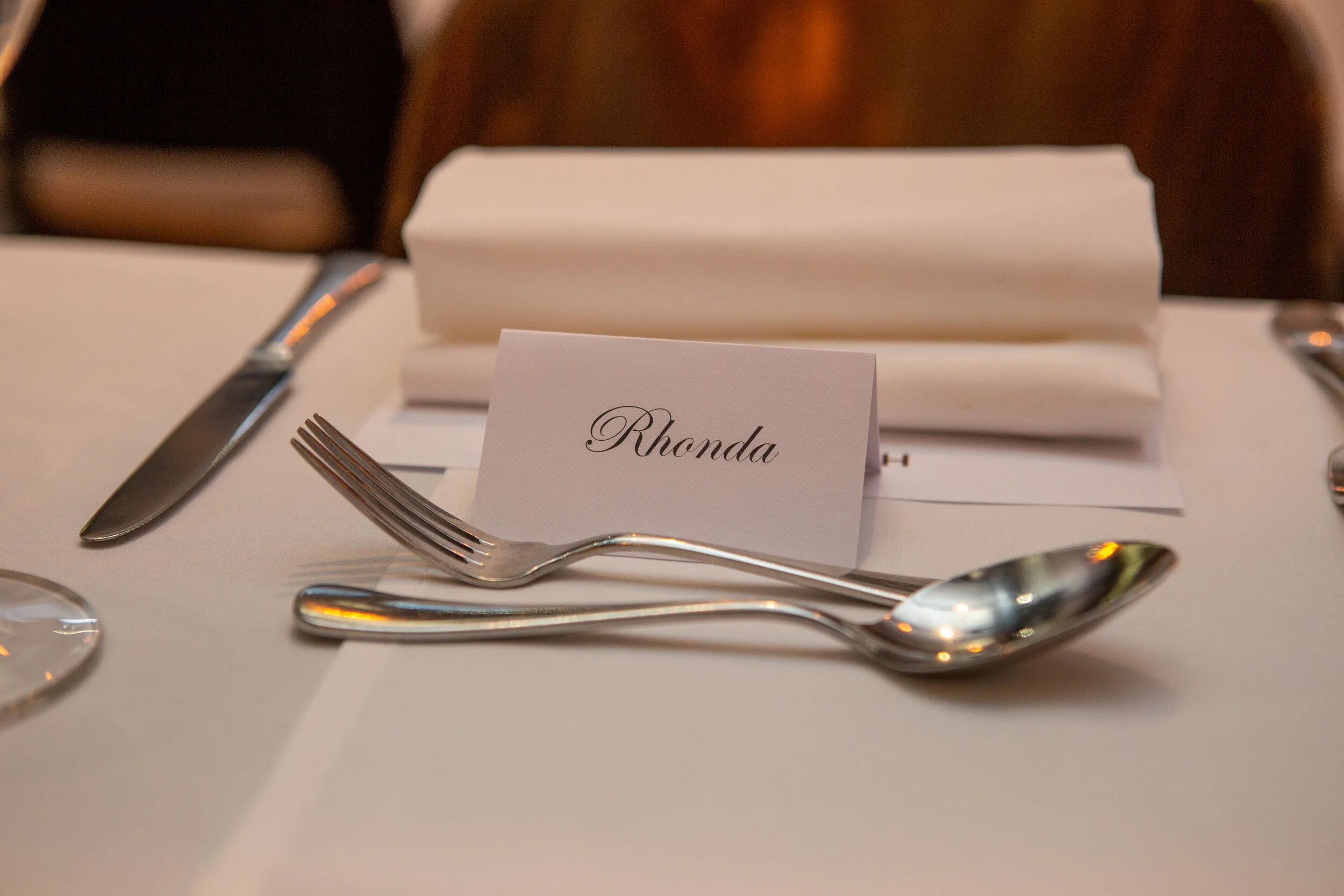 Table setting with a name card labeled Rhonda, a fork, a spoon, a knife, a folded napkin, and an empty plate on a white tablecloth.