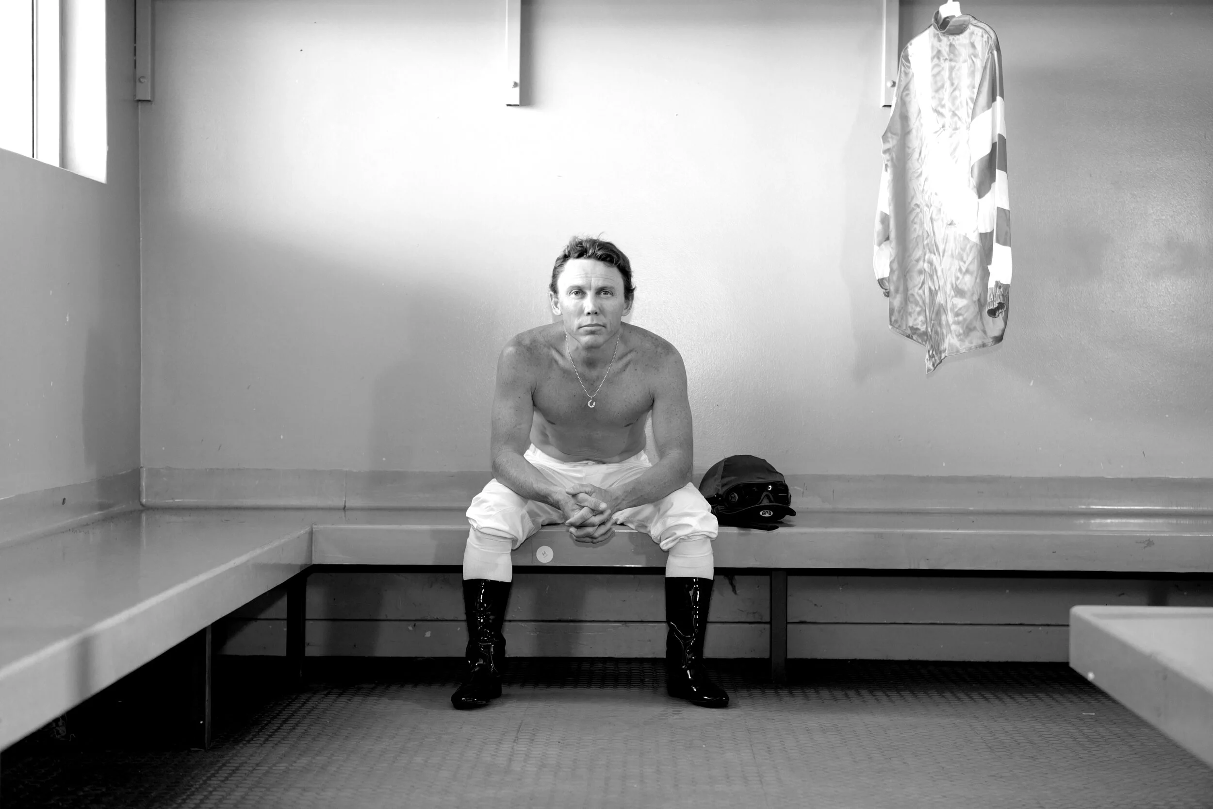 A shirtless man sitting on a bench in a locker room, wearing white pants and black rain boots, with a cap placed next to him and a hanging garment on the wall behind.