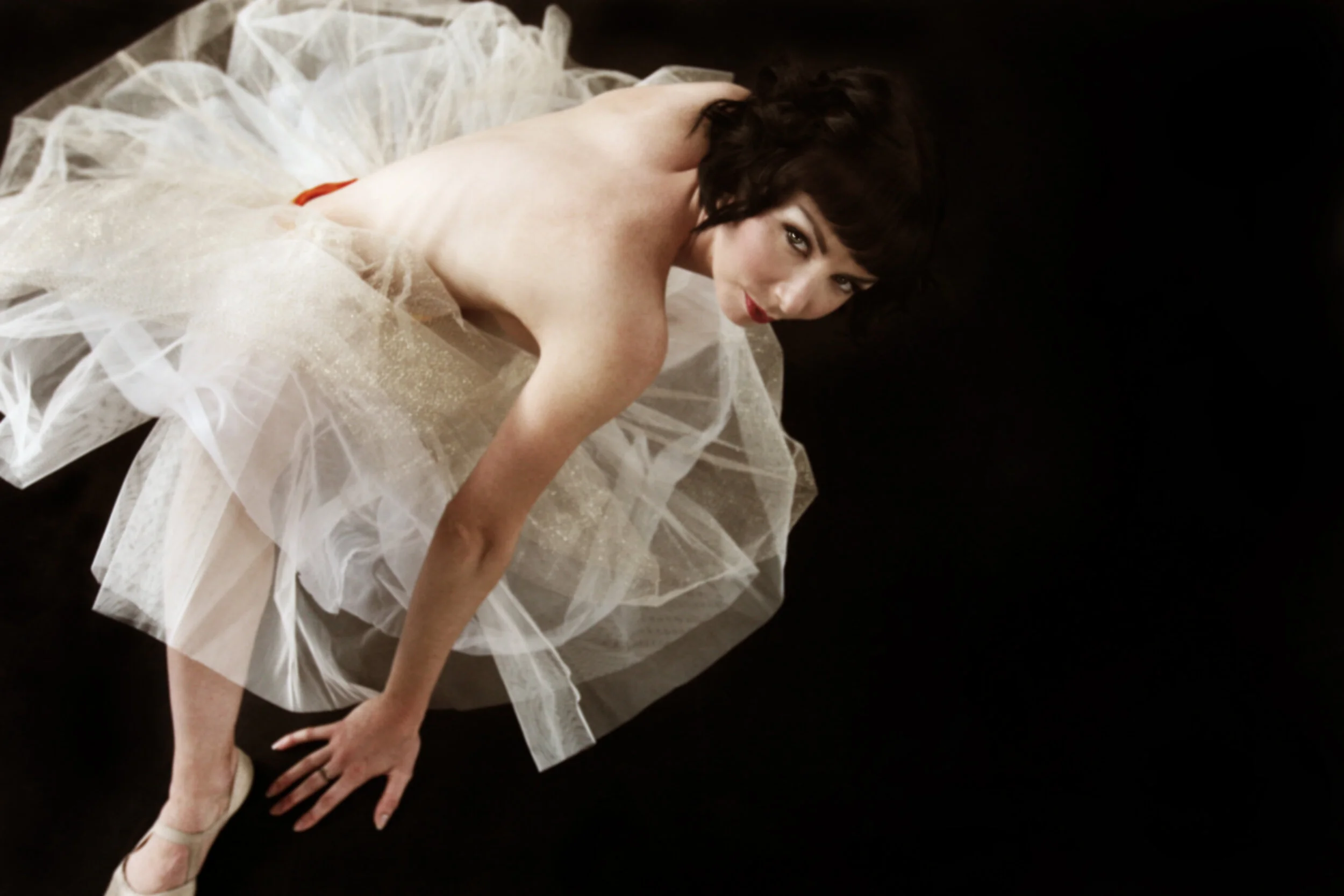 A woman in a white, sparkly tulle dress with black hair and red lipstick, lying on a dark surface and looking at the camera.