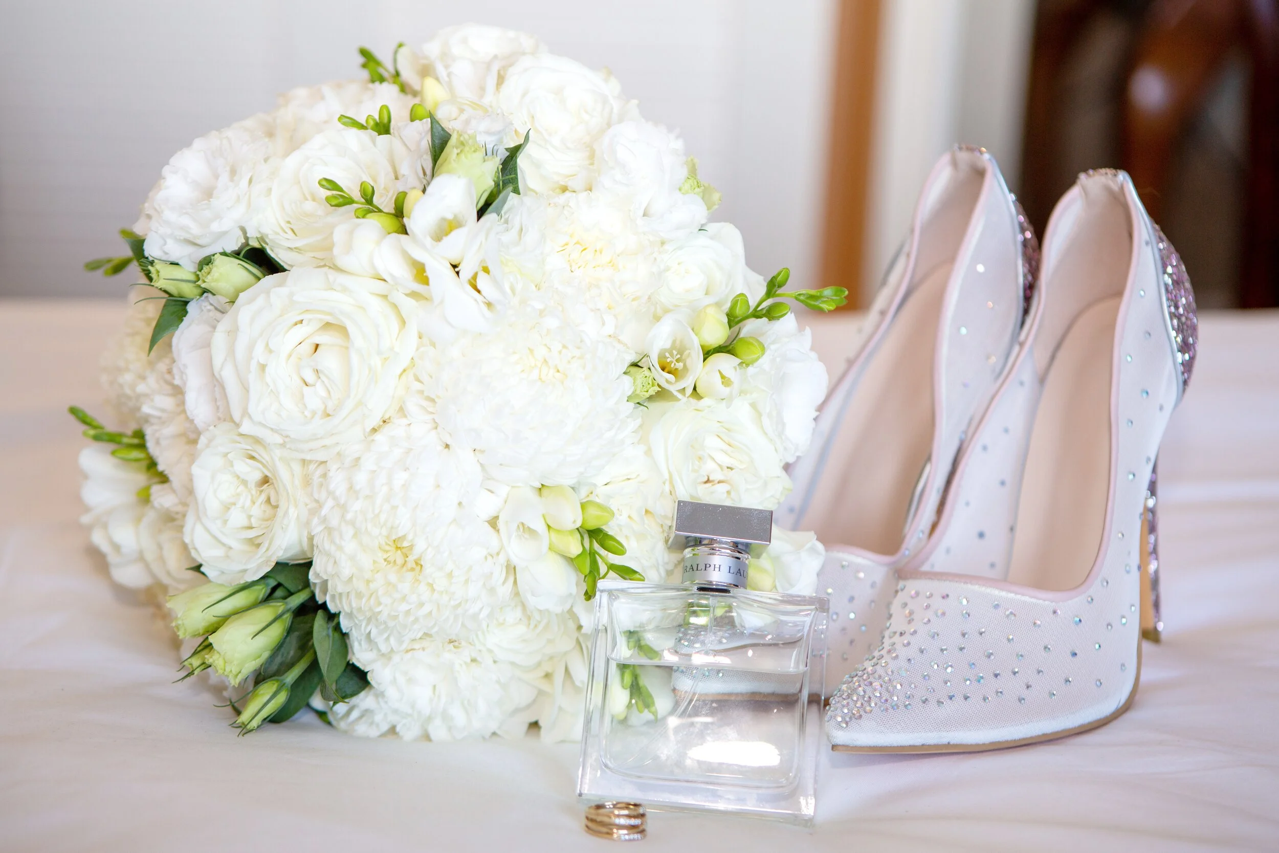 Wedding bouquet, white high-heeled shoes with rhinestones, a perfume bottle, and wedding rings on a table.