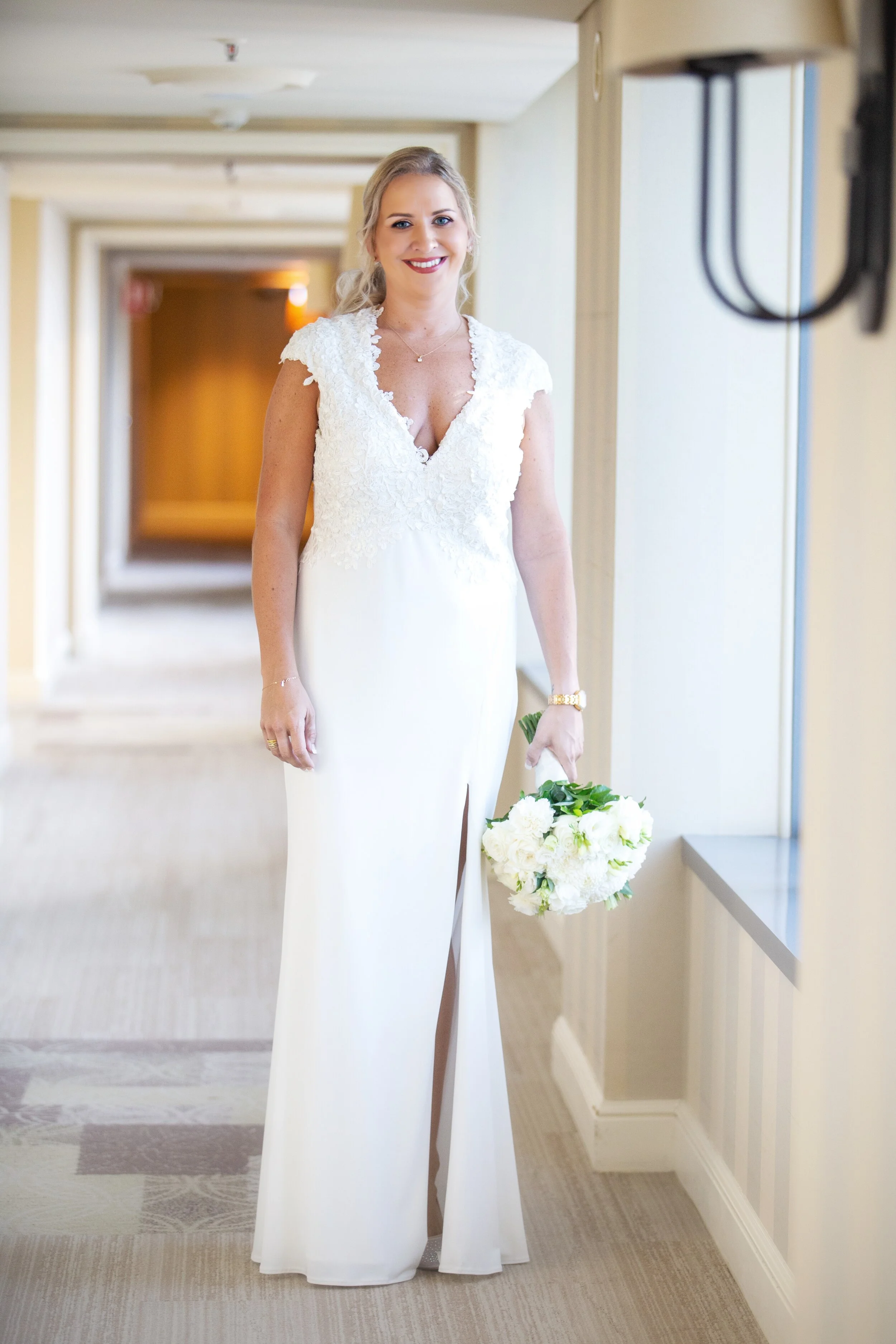 A bride in a white wedding gown holding a bouquet of white flowers, standing in a hallway, smiling.