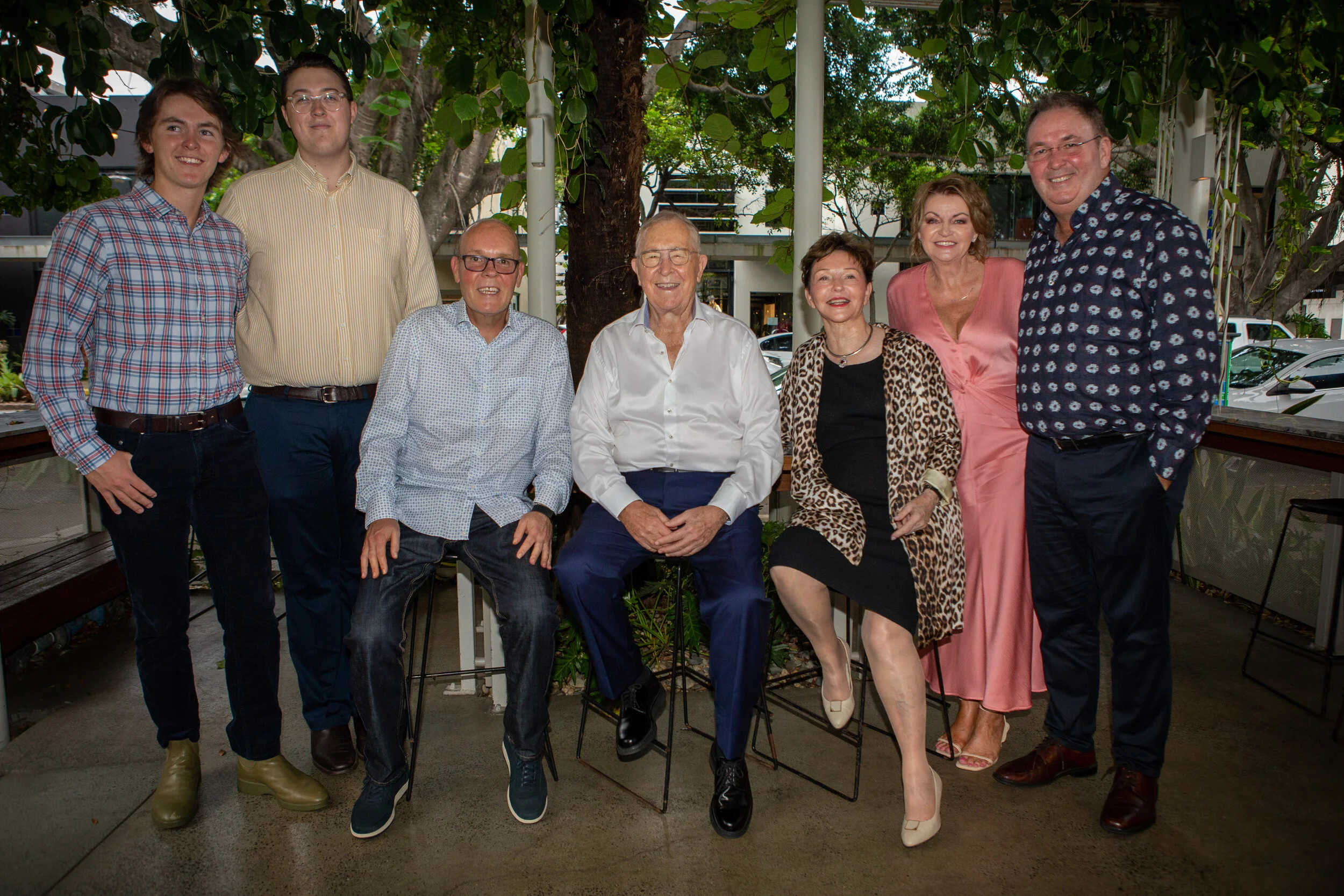 Group of nine people, six women and three men, smiling and posing for a photo on a patio with trees and parked cars in the background.