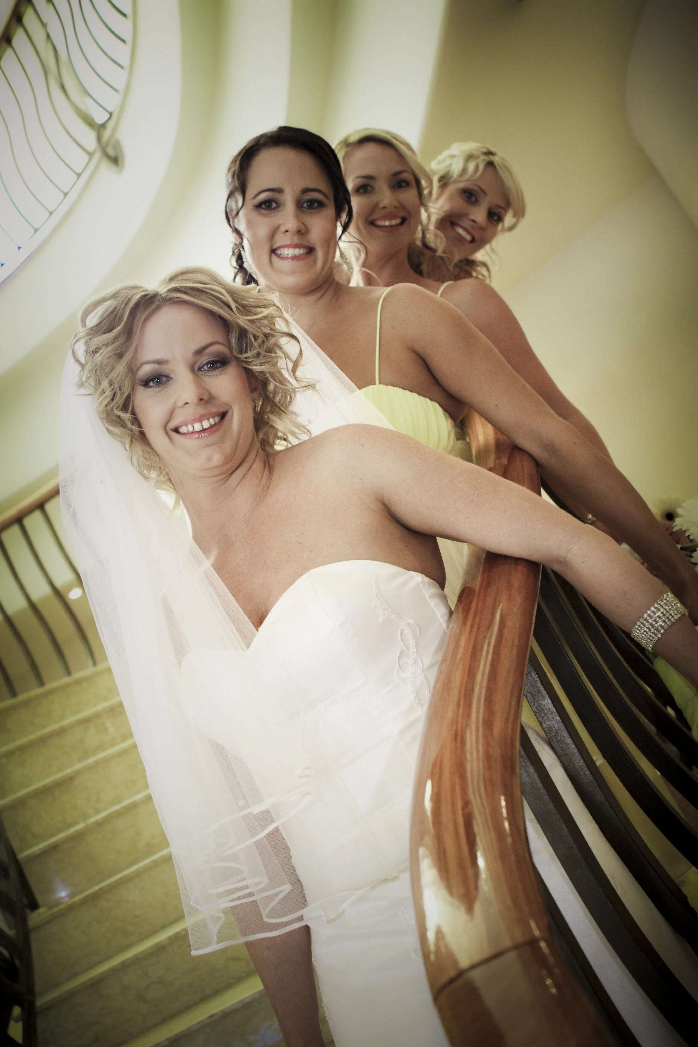 Four women, including a bride in a white wedding dress with a veil, are standing on a staircase, smiling for the photo.