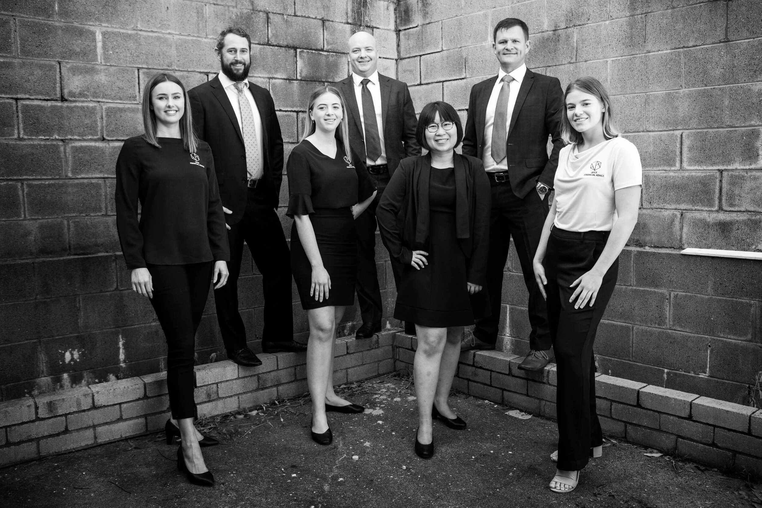 Group of eight professionally dressed people standing together outdoors, positioned against brick walls, smiling at the camera.