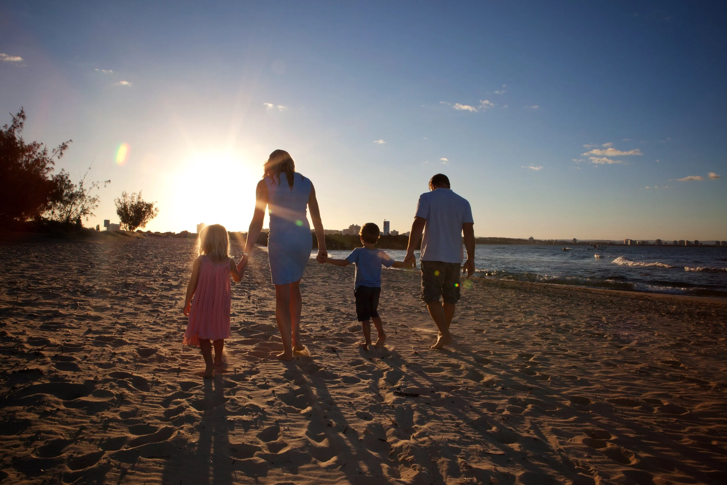 Family walking on the beach at sunset, holding hands, with the ocean on the right side and a city skyline in the background.