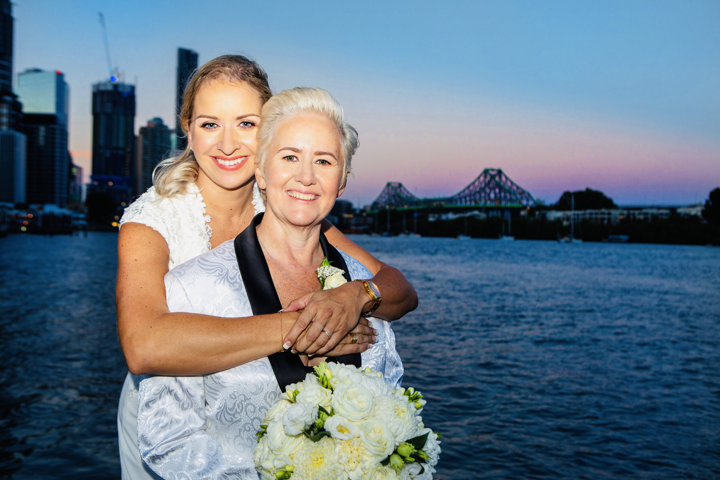A woman with short gray hair and a woman with long blond hair smiling and embracing near a body of water at sunset, with city skyline and a bridge in the background.