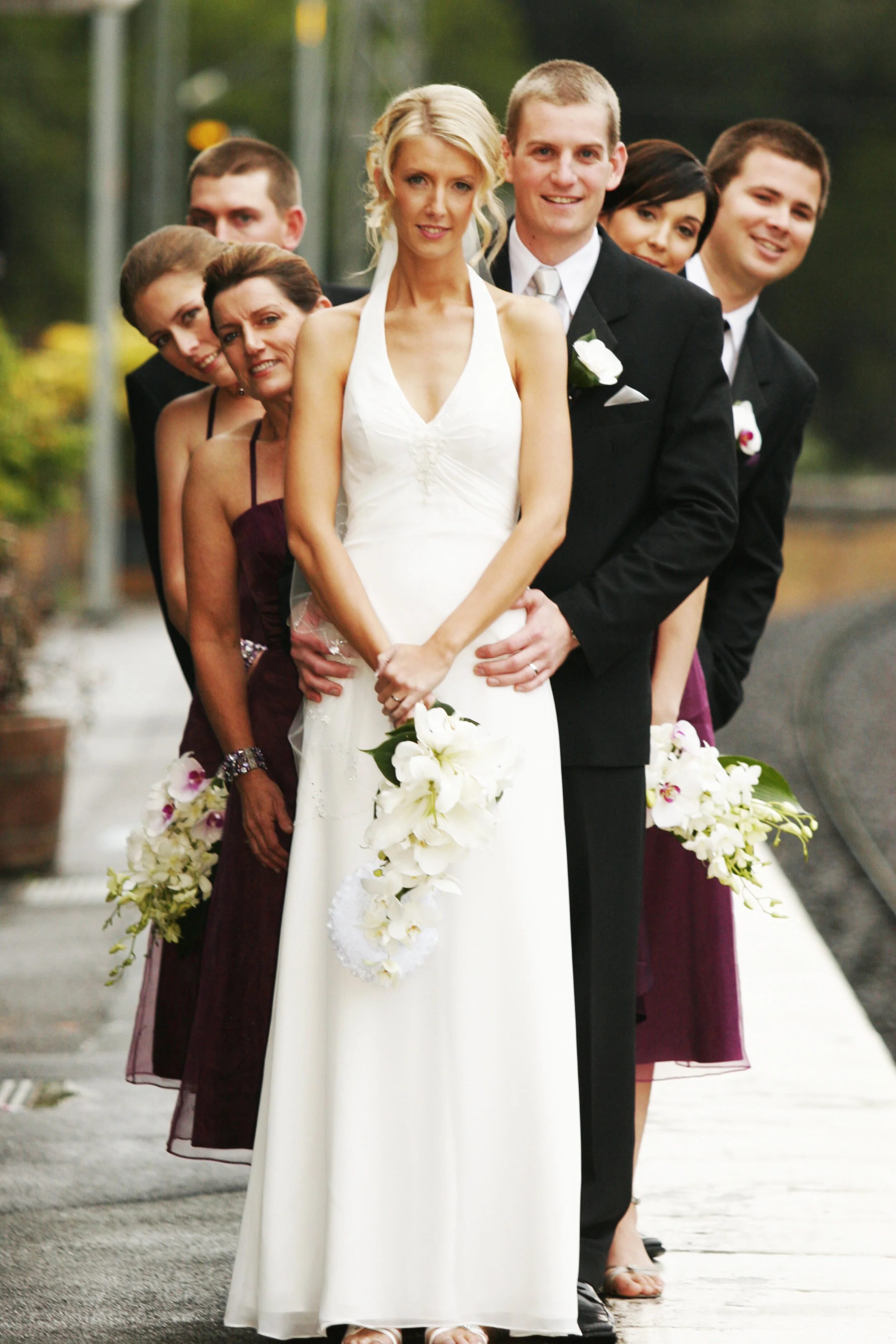 A wedding party standing in a line on a sidewalk, including a bride in a white wedding dress holding a floral bouquet, a groom in a black tuxedo, and four women and men wearing formal attire.