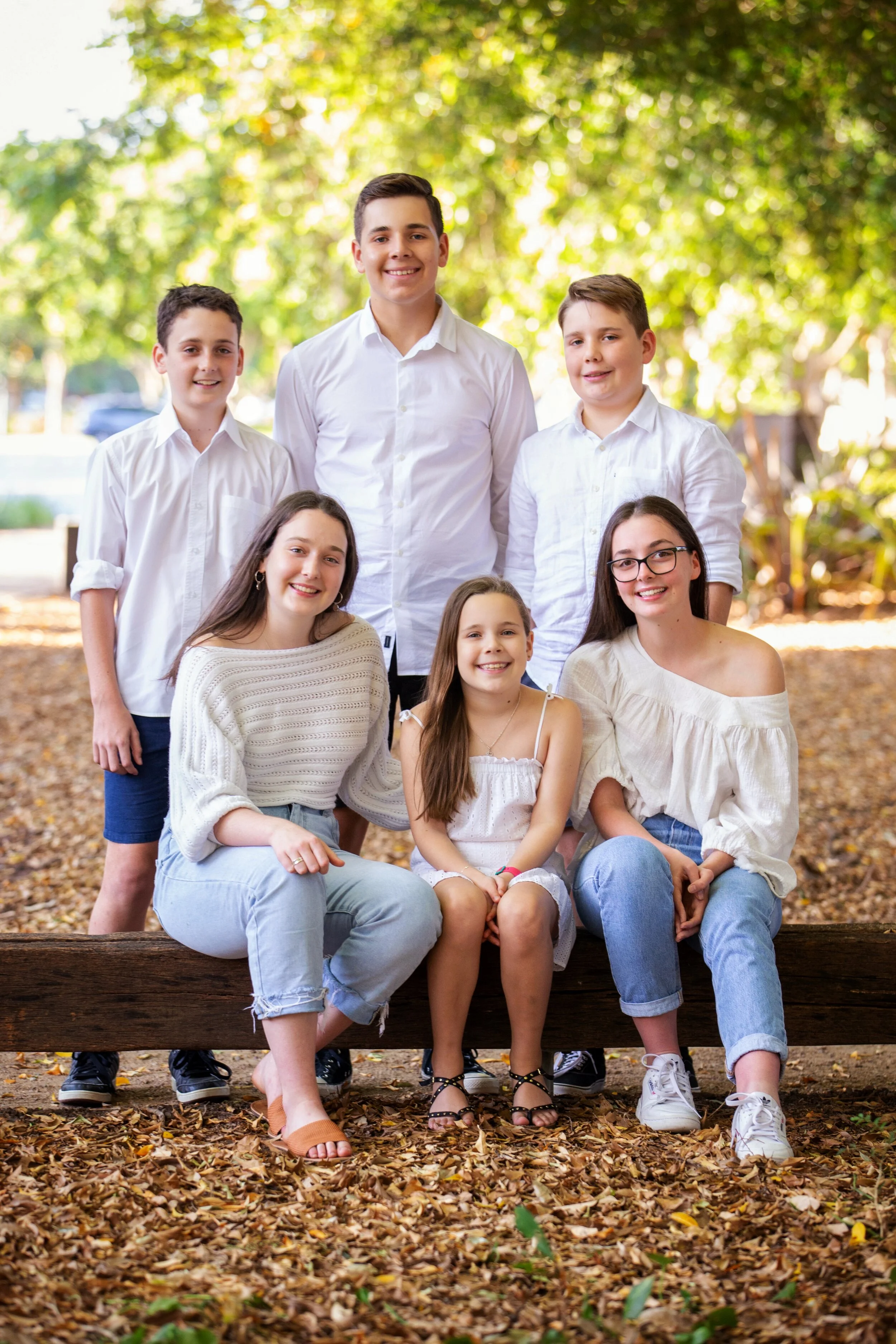 Group of seven children, four boys and three girls, smiling outdoors on a sunny day with fallen leaves and green trees in the background.