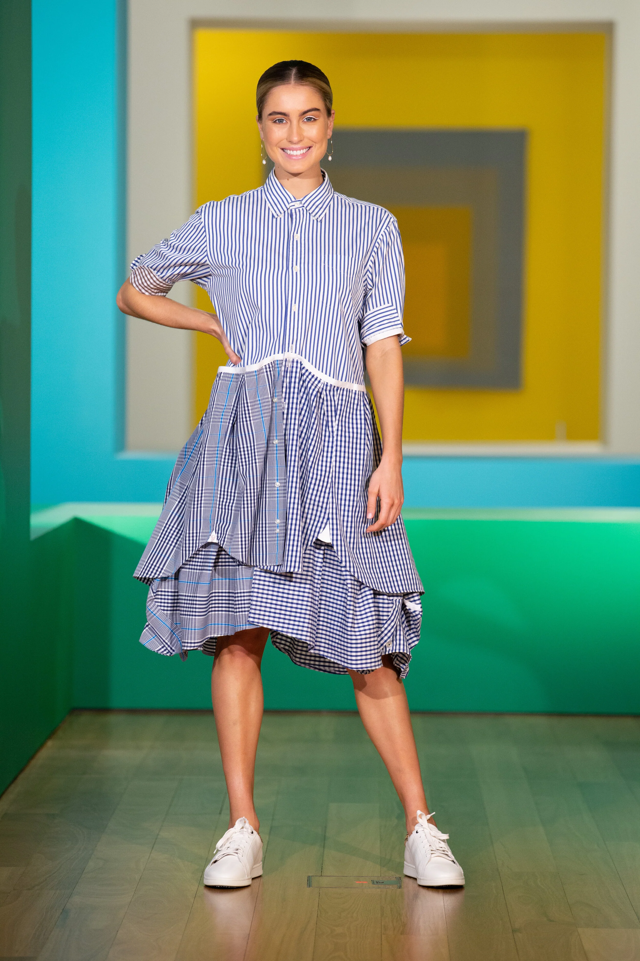 A woman wearing a blue and white striped dress with sneakers, standing indoors in front of a colorful geometric background, smiling at the camera.