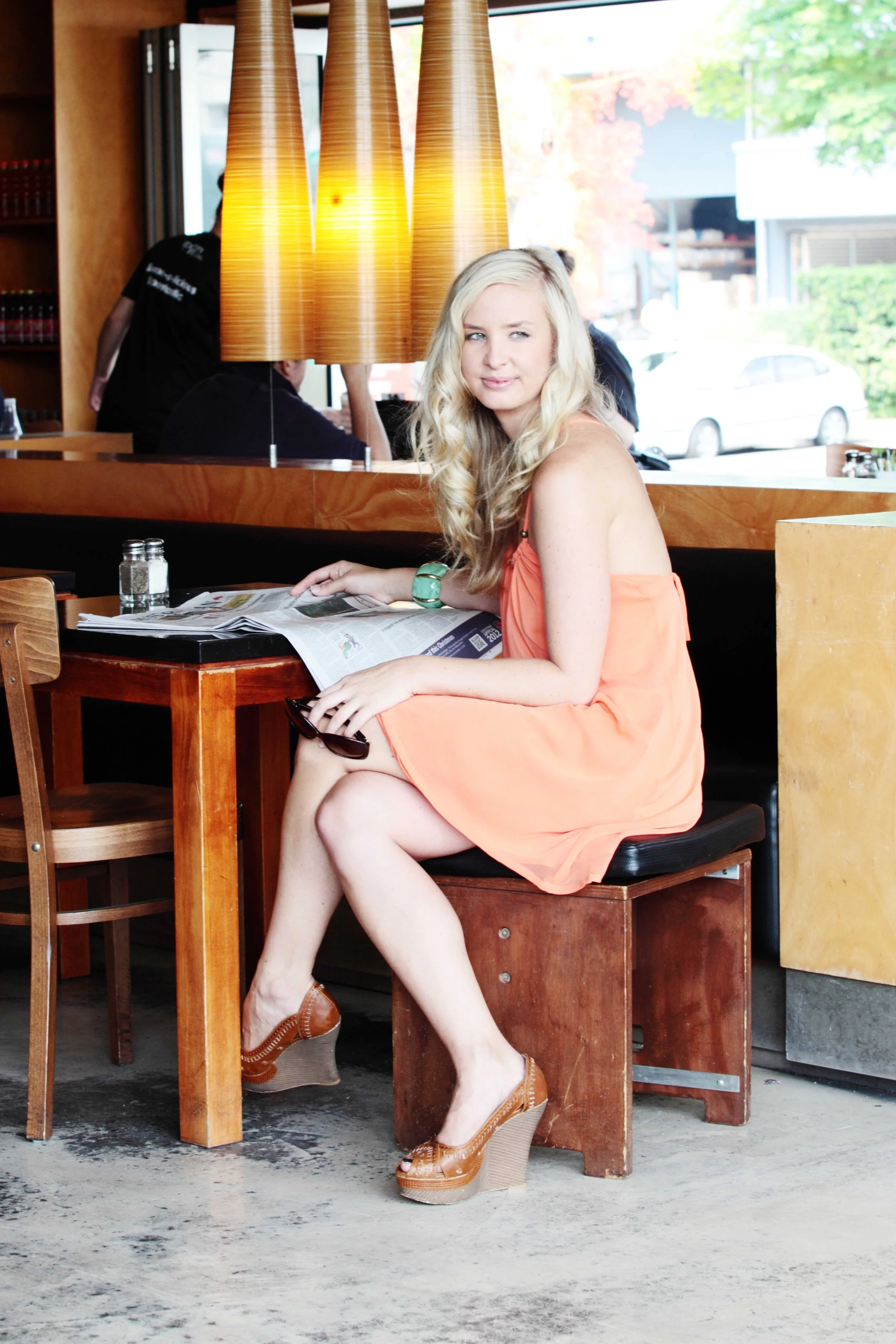 A woman in a peach dress and tan wedge shoes sitting in a restaurant, holding a newspaper and a pair of sunglasses, with warm pendant lighting overhead and a large window showing parked cars outside.