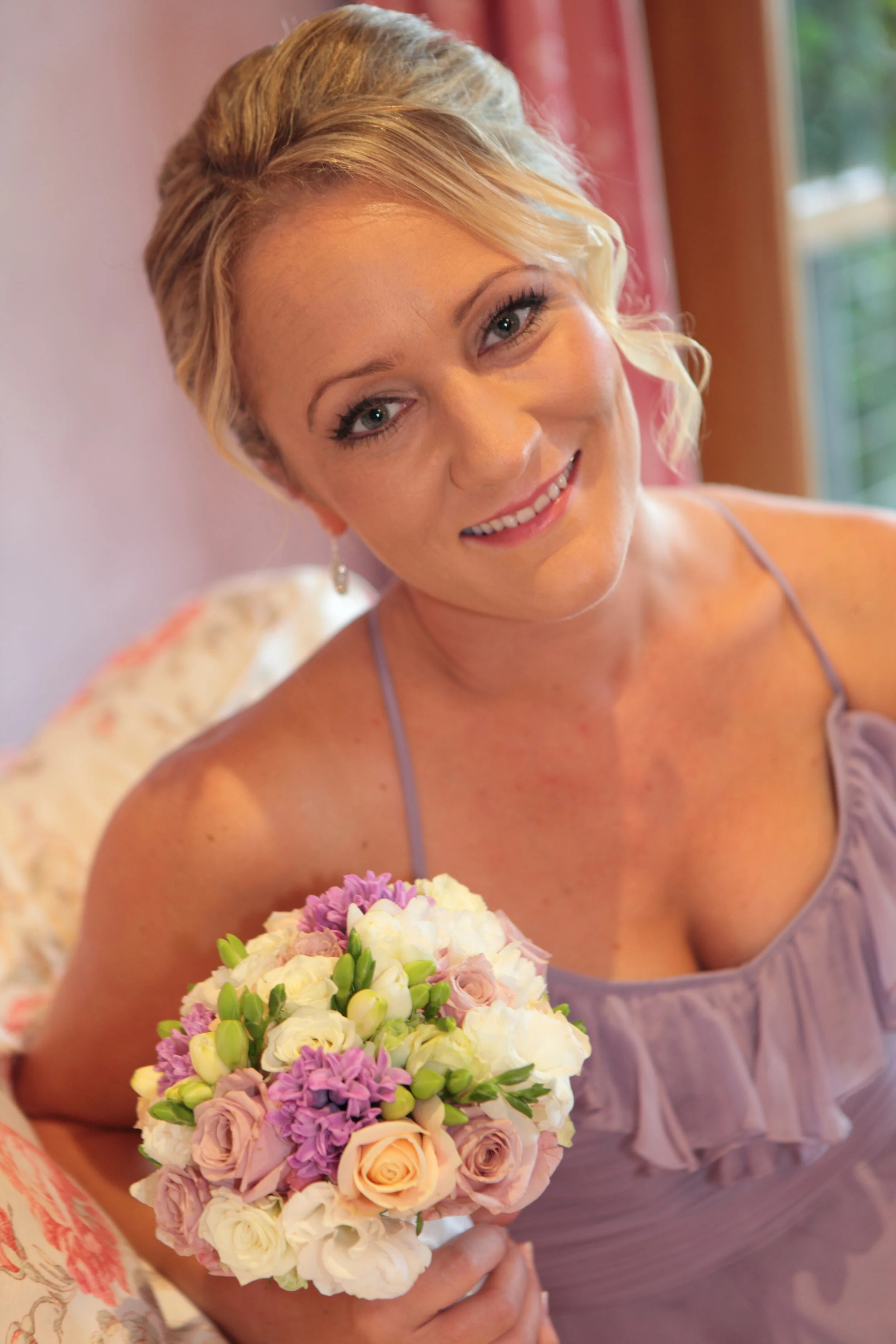 A woman in a lavender dress holding a bouquet of flowers and smiling at the camera.