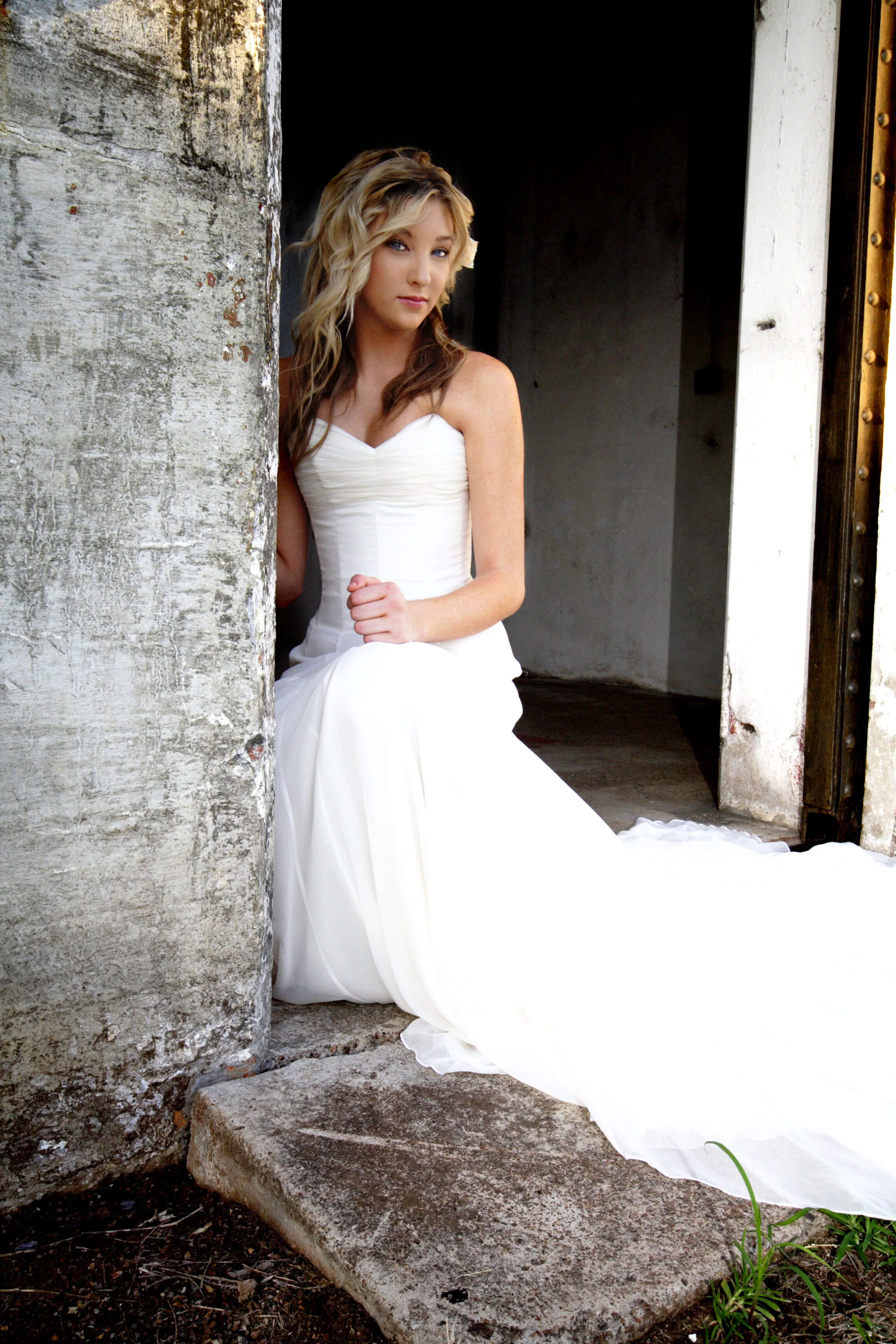 A woman in a white wedding dress sitting on a step outside a rustic building, looking at the camera.