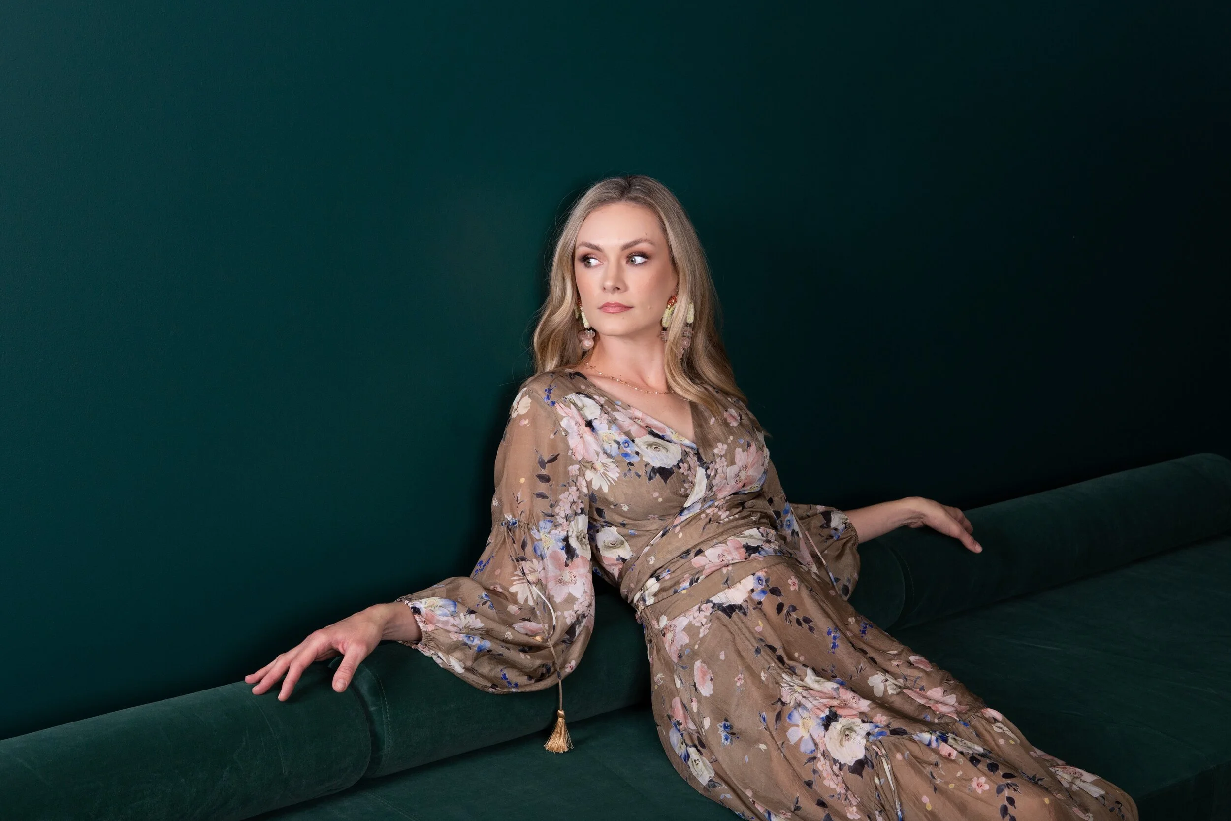 A woman with long blonde hair wearing a floral dress sits on a dark green couch against a matching dark green wall, looking to her left.