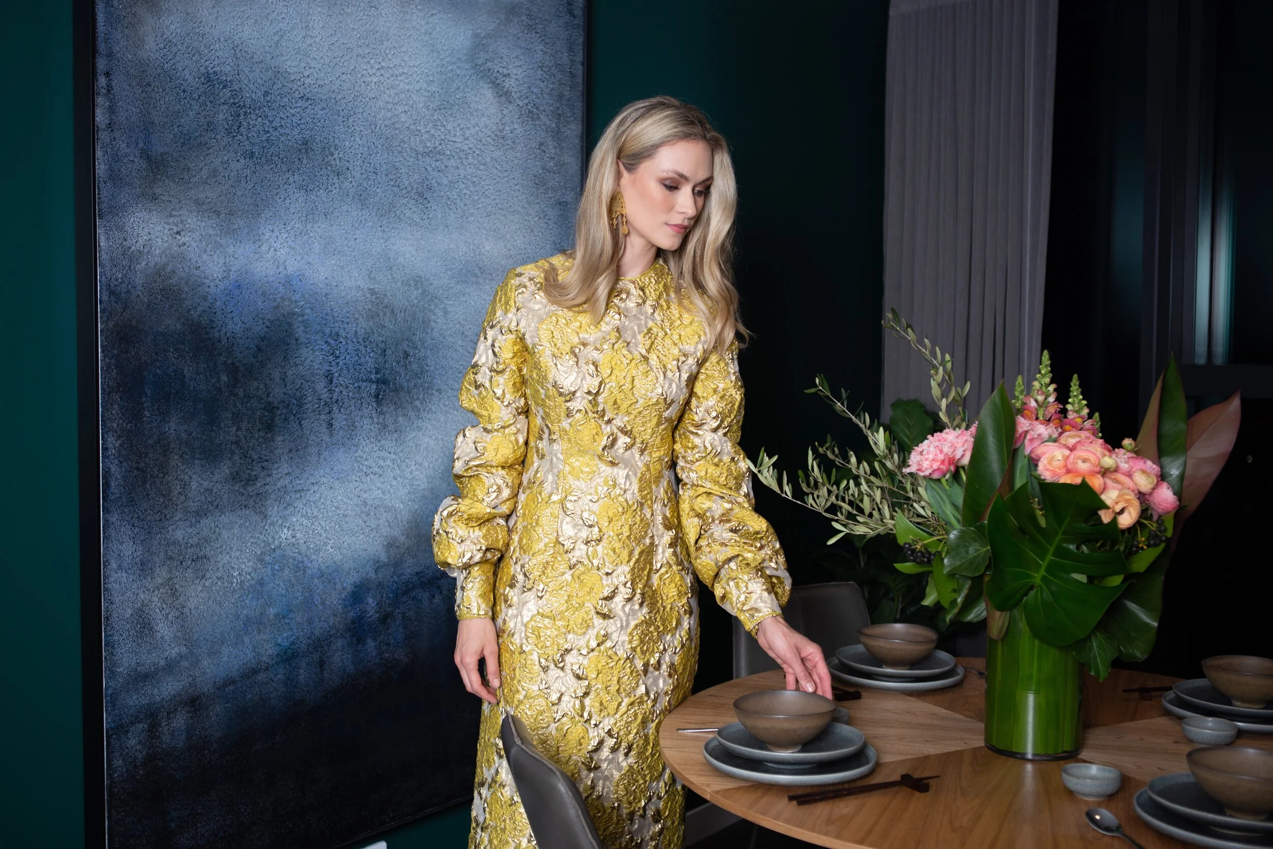 Woman in a gold floral dress setting a dinner table with a large floral arrangement in a green vase.