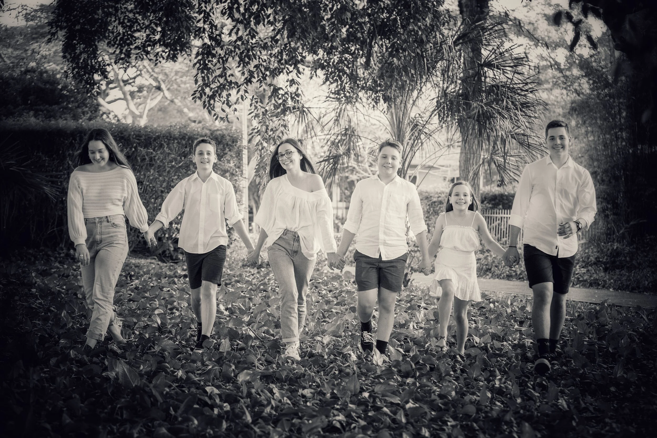 A family of seven holding hands and walking through a wooded outdoor area. All are smiling and casually dressed in light-colored clothing.
