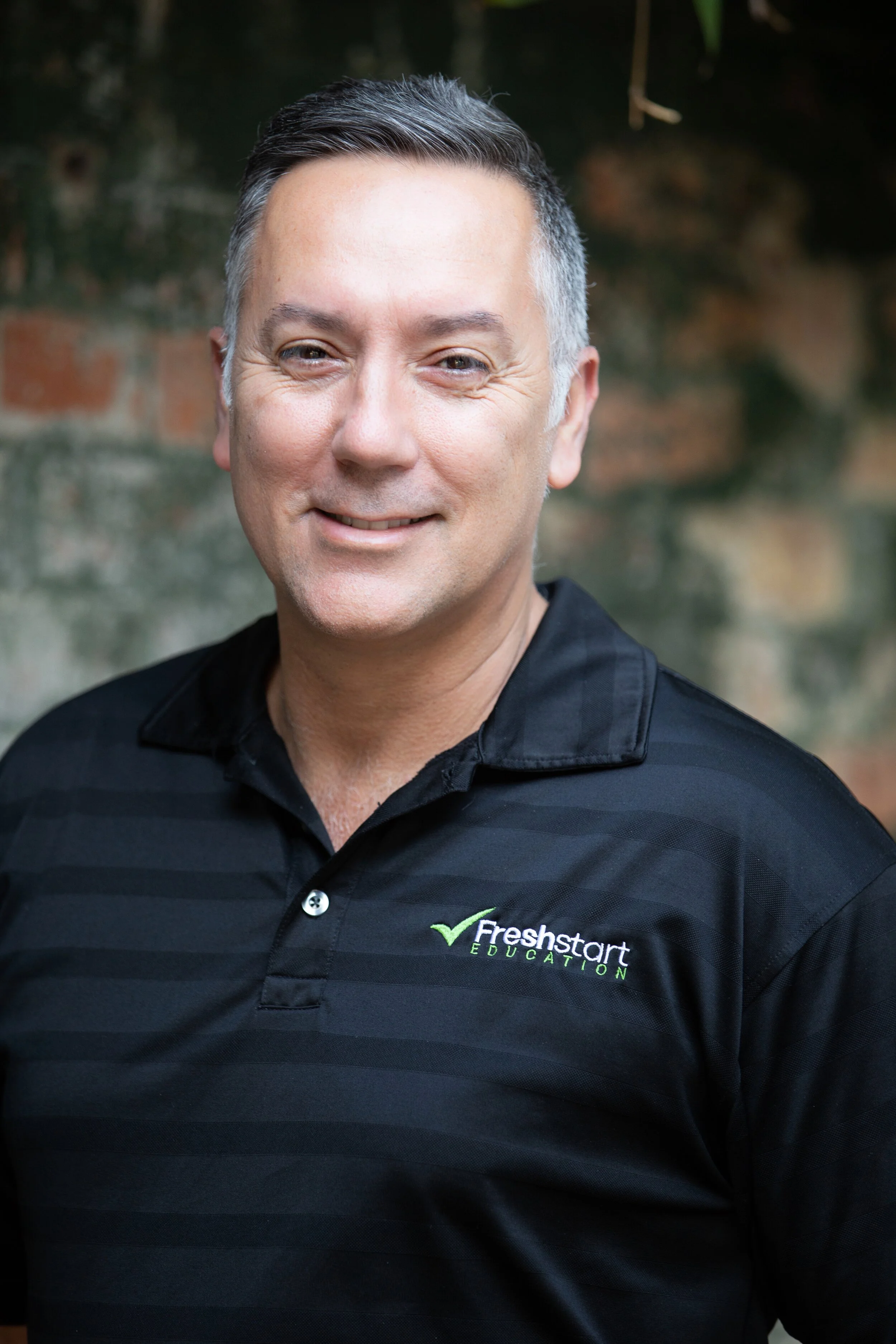 A man smiling, wearing a black polo shirt with a logo that reads 'Freshstart EDUCATION,' standing outdoors in front of a blurred brick and green plant background.