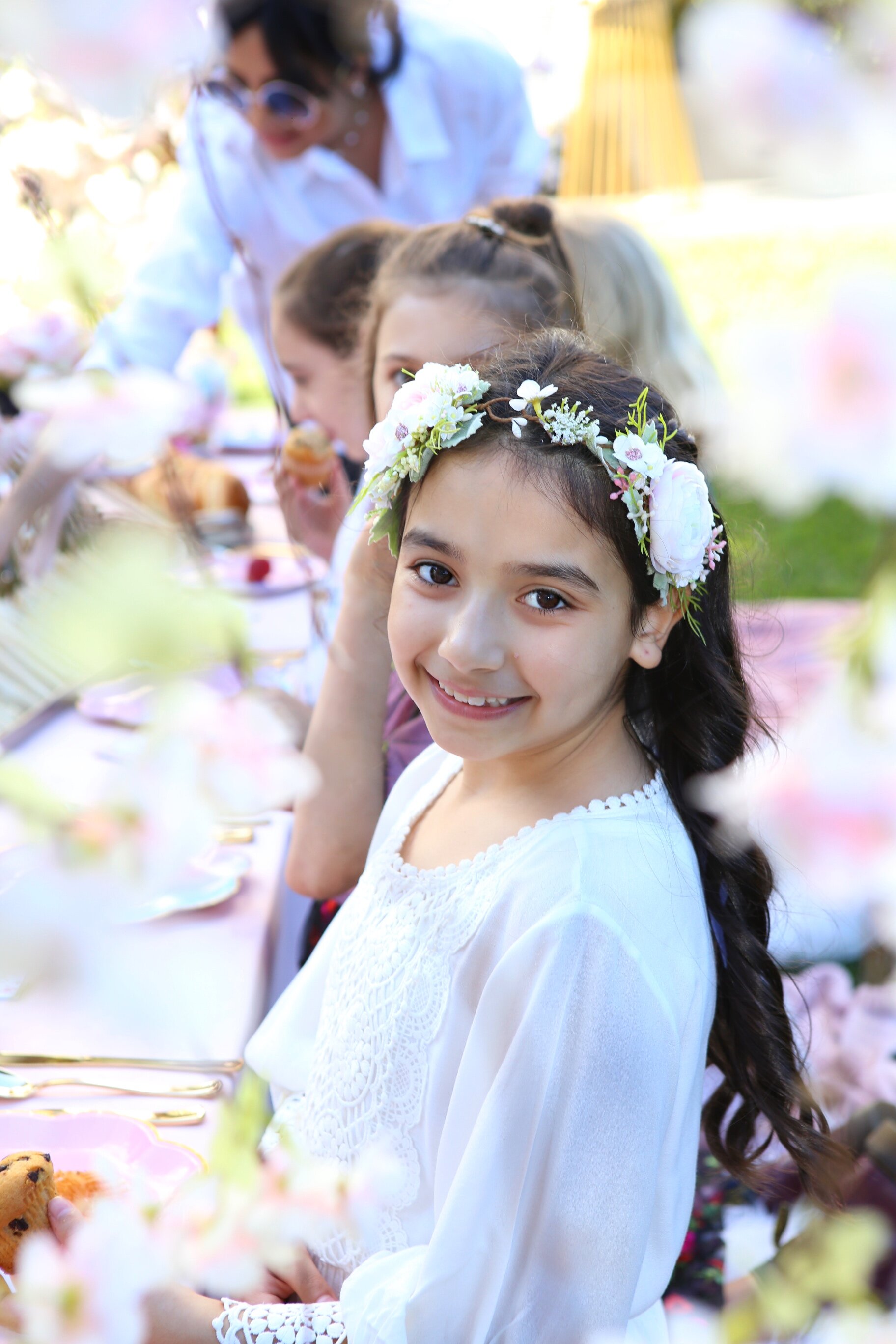 A young girl with dark hair wearing a floral headband and white dress, smiling at a fairy tale-themed outdoor tea party with women and children in the background.