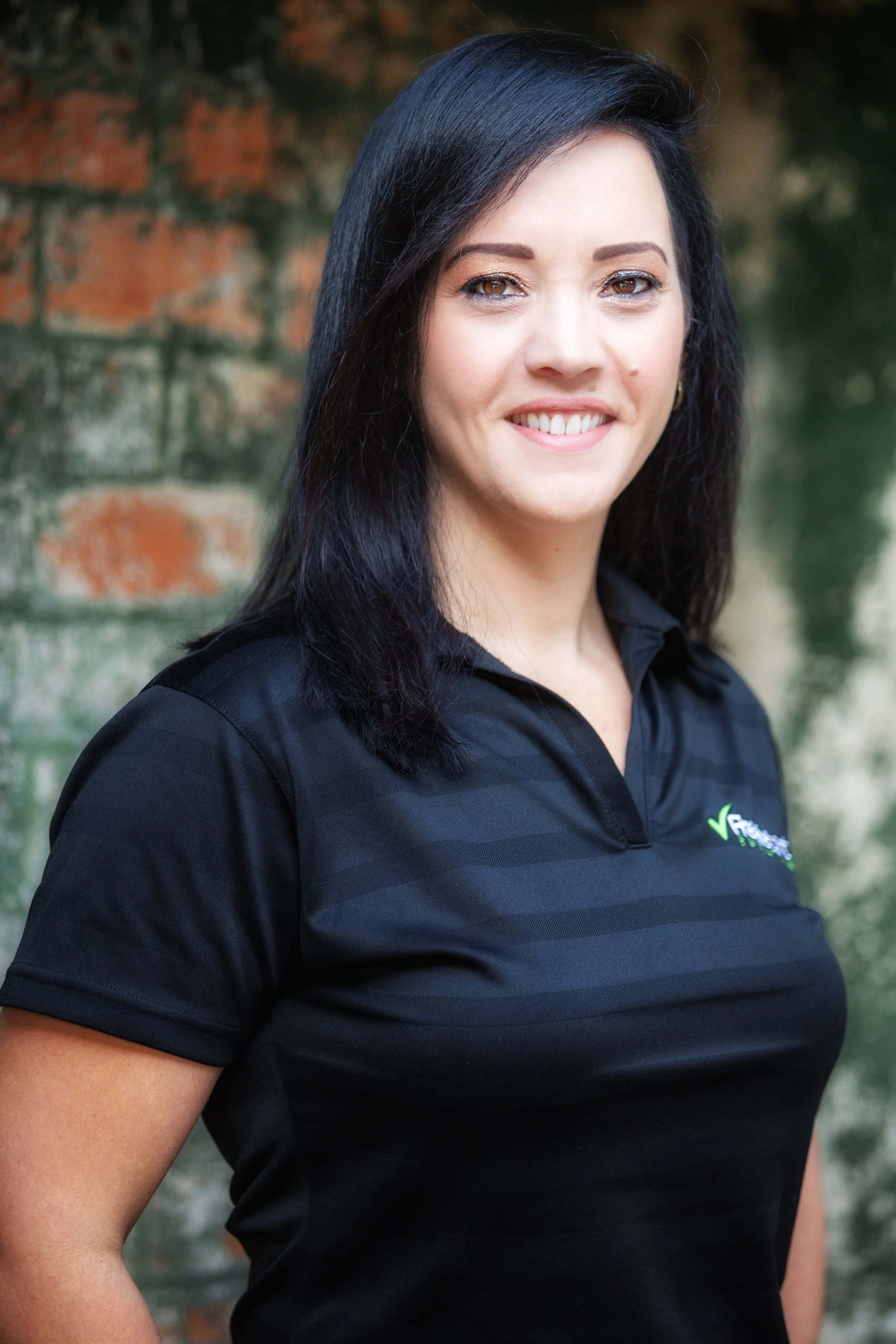 A woman with shoulder-length black hair, wearing a black athletic polo shirt, standing outdoors with a brick wall background, smiling at the camera.