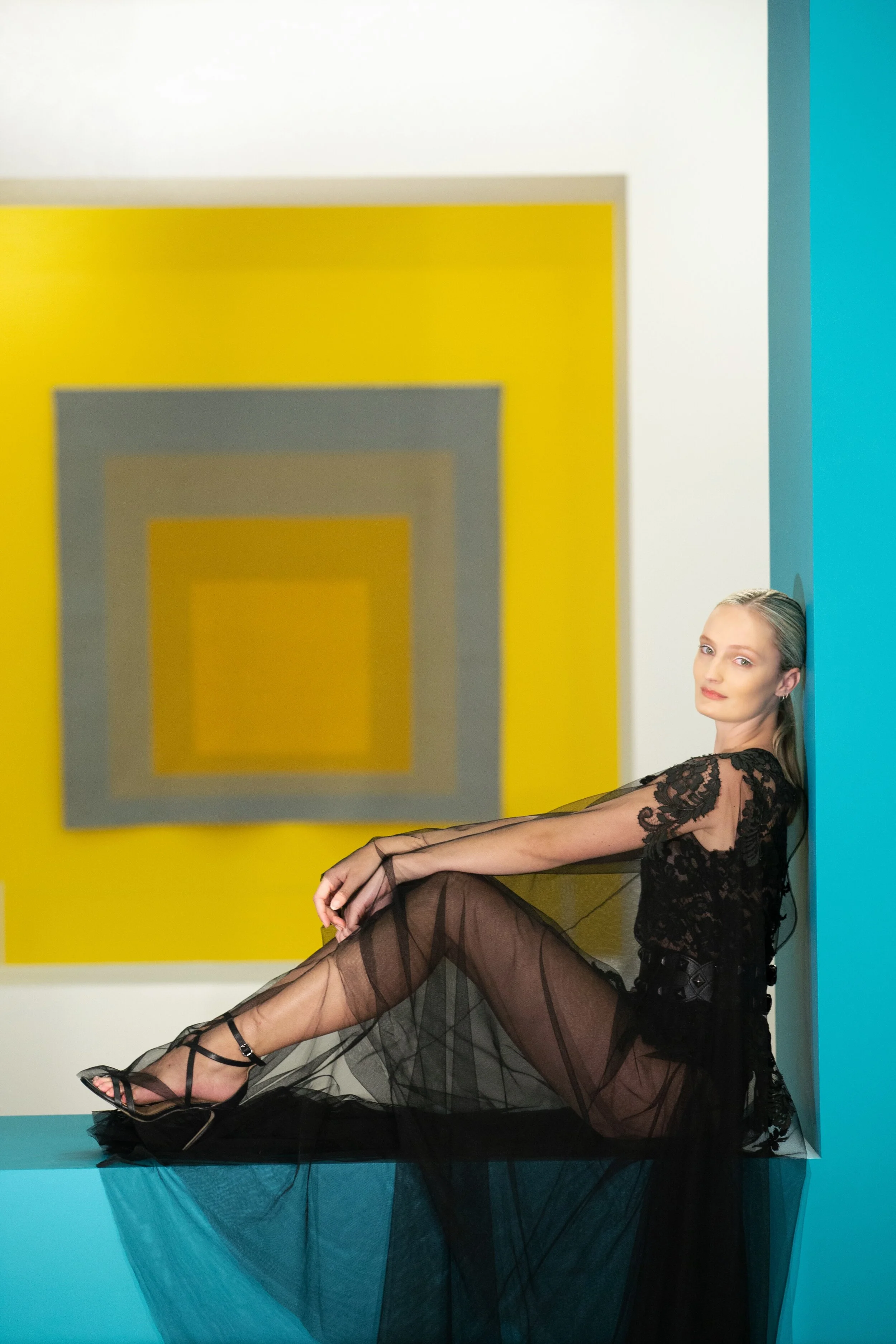 A woman in a black lace dress and high heels sitting on a blue ledge against a turquoise wall, with abstract yellow and gray square artwork in the background.
