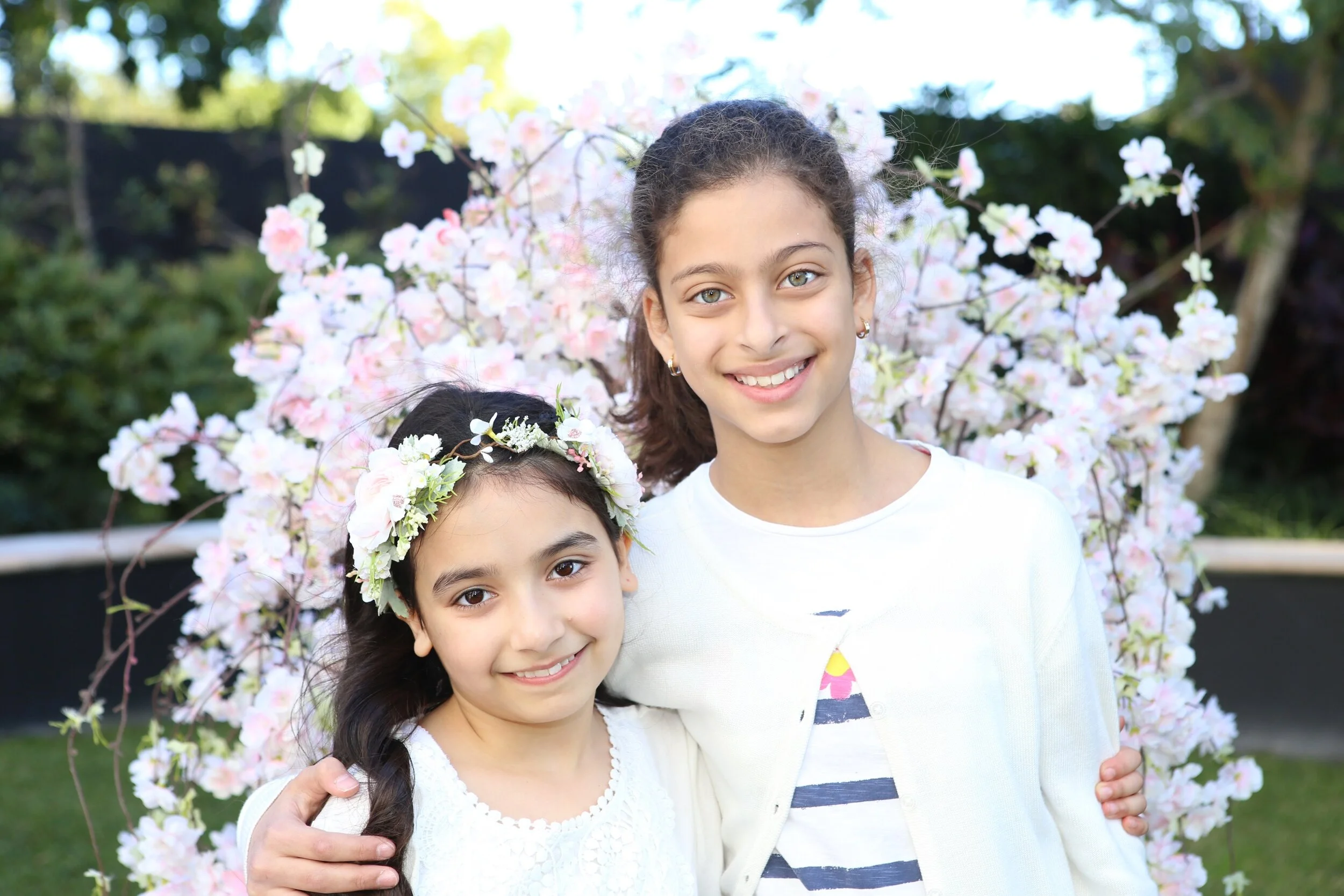 Two young girls standing outdoors in front of a pink flowering tree, smiling with their arms around each other.
