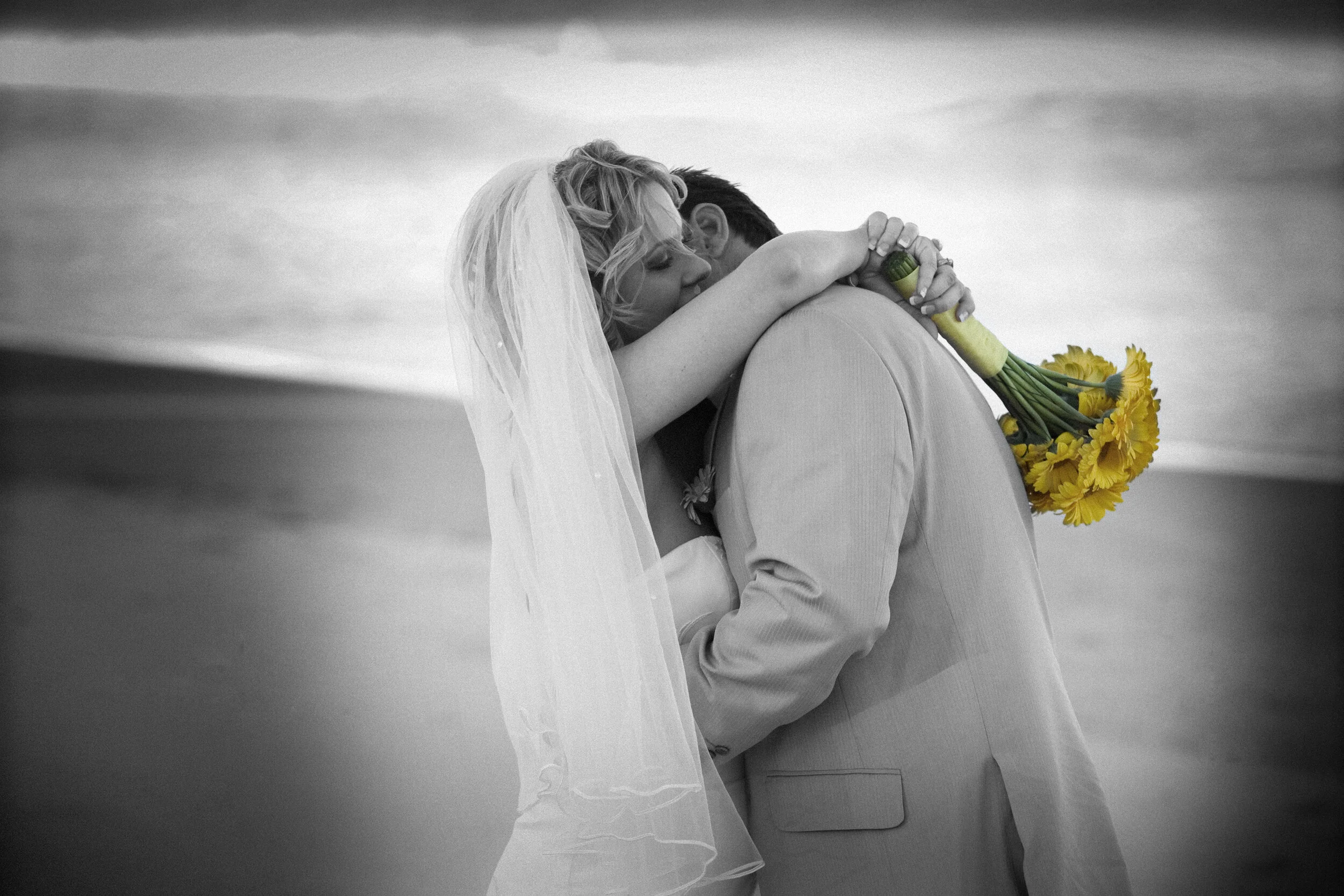 A bride and groom embracing on a beach, with the bride holding a bouquet of yellow flowers.